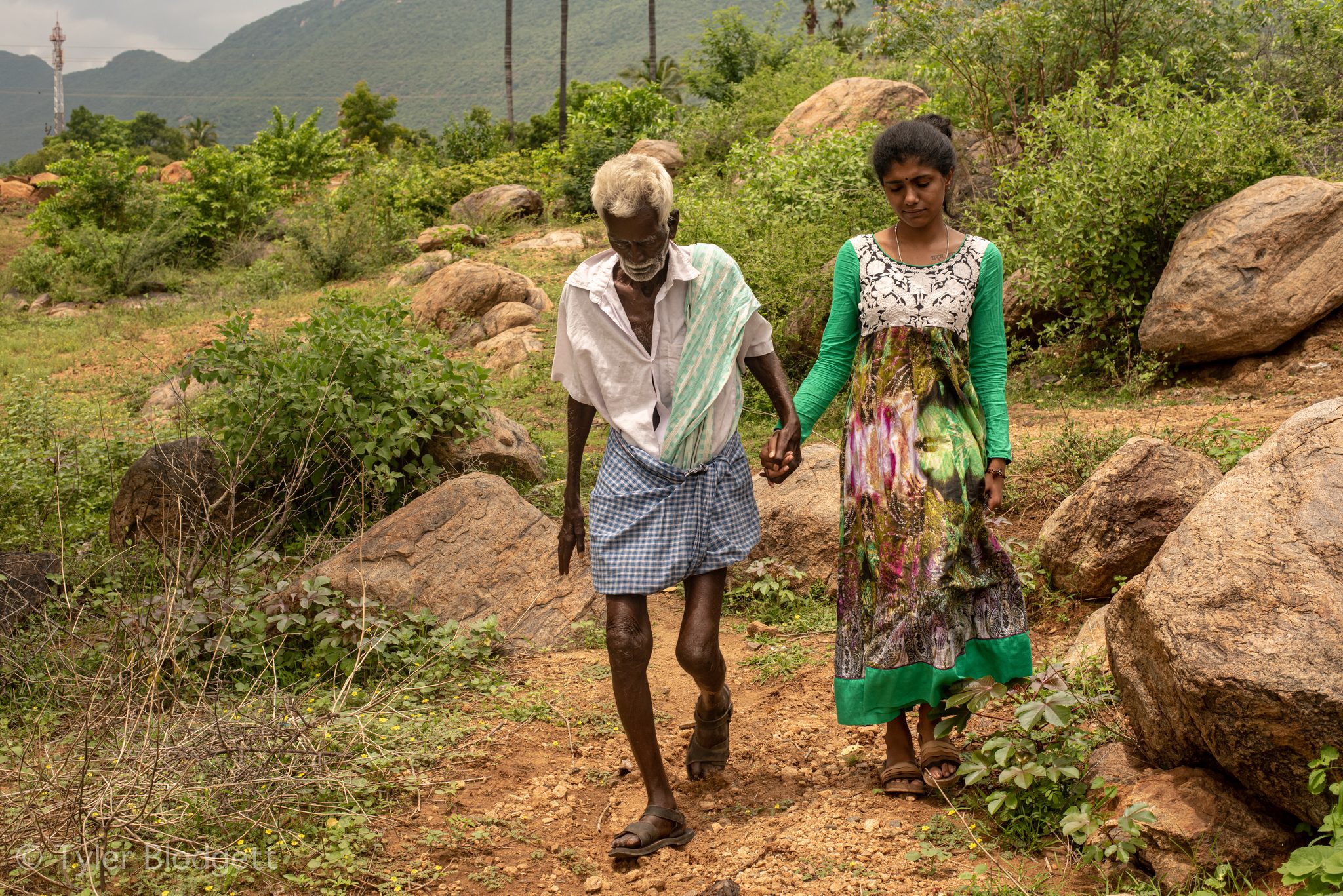 The elder walks hand in hand with a young woman along a rocky path.