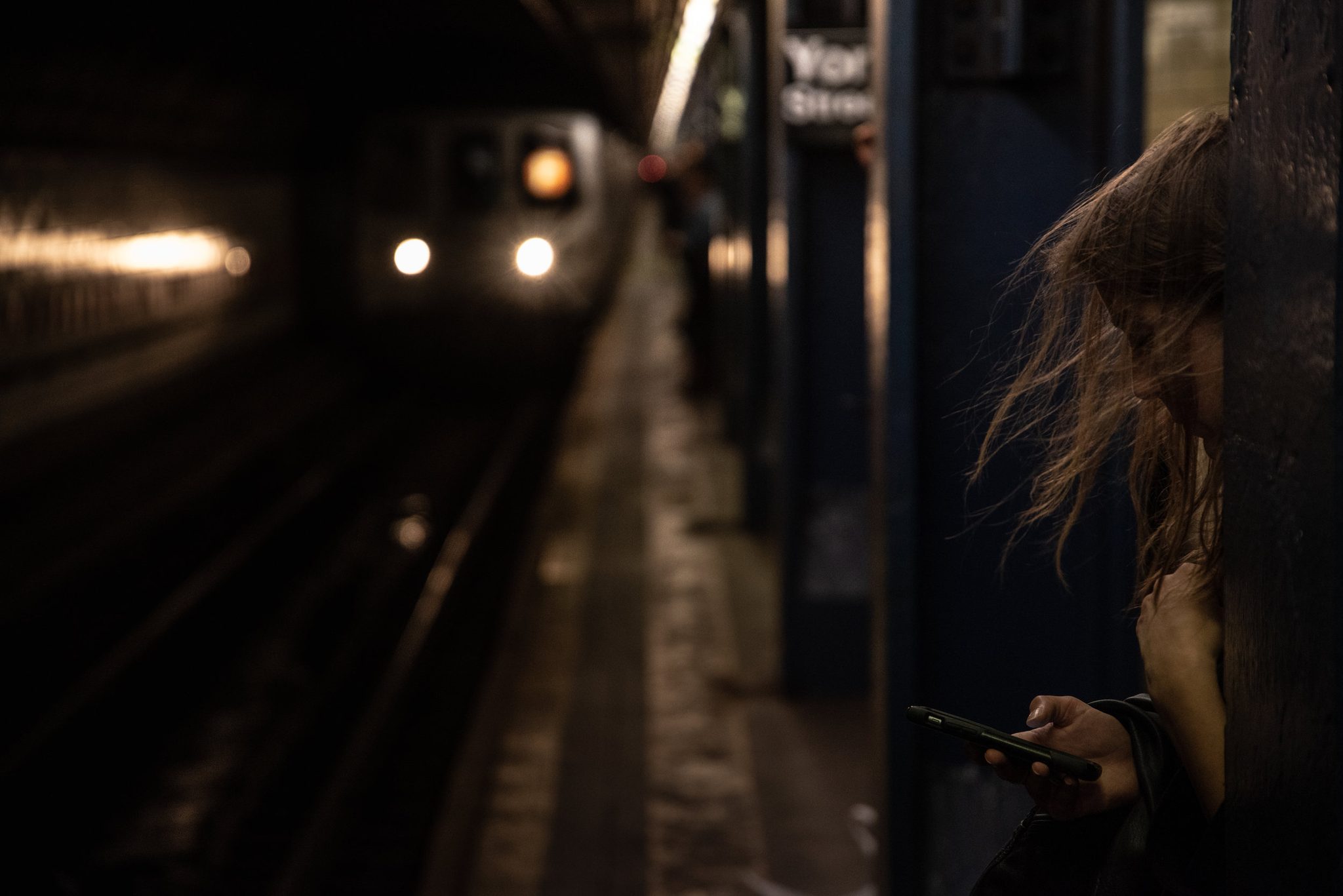 Headlights swell in the tunnel while a woman on the platform keeps her eyes on her phone.