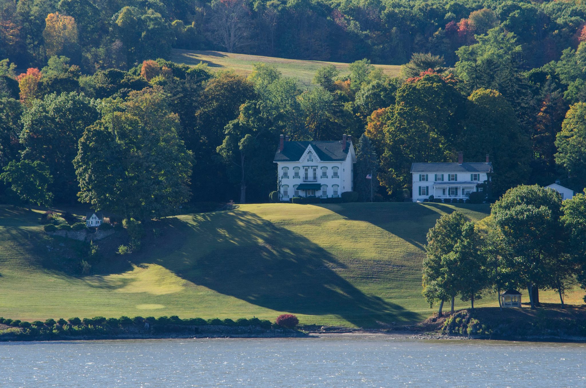 A white house sits above the river on a green hill, the trees just starting to turn.