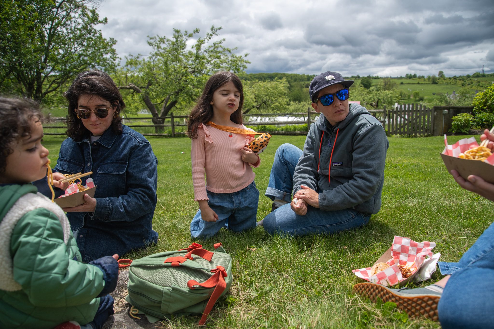 A family picnics on the grass, paper boats of lunch passed around.