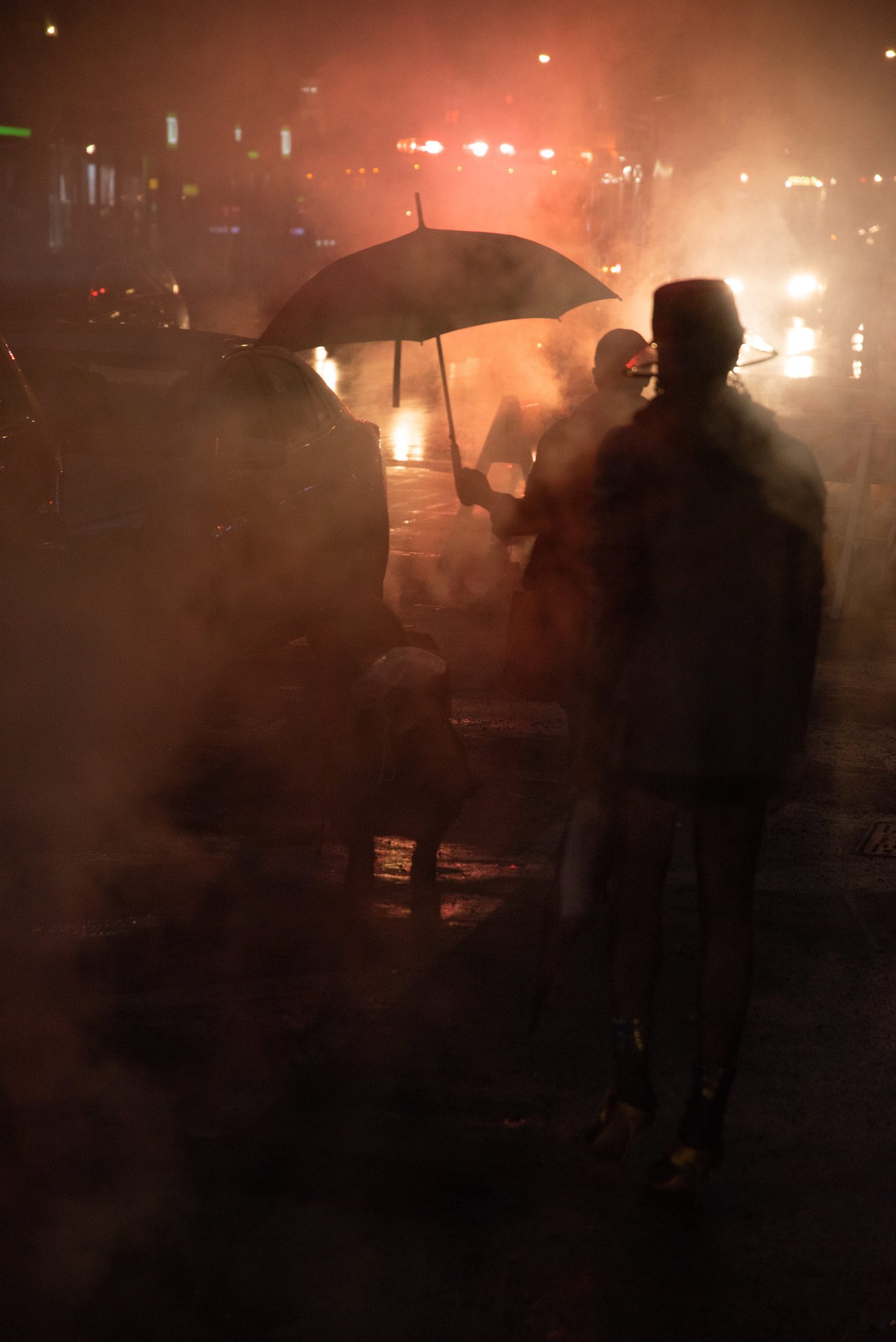 Shadows drift under umbrellas through the steam and the red glow of the street.