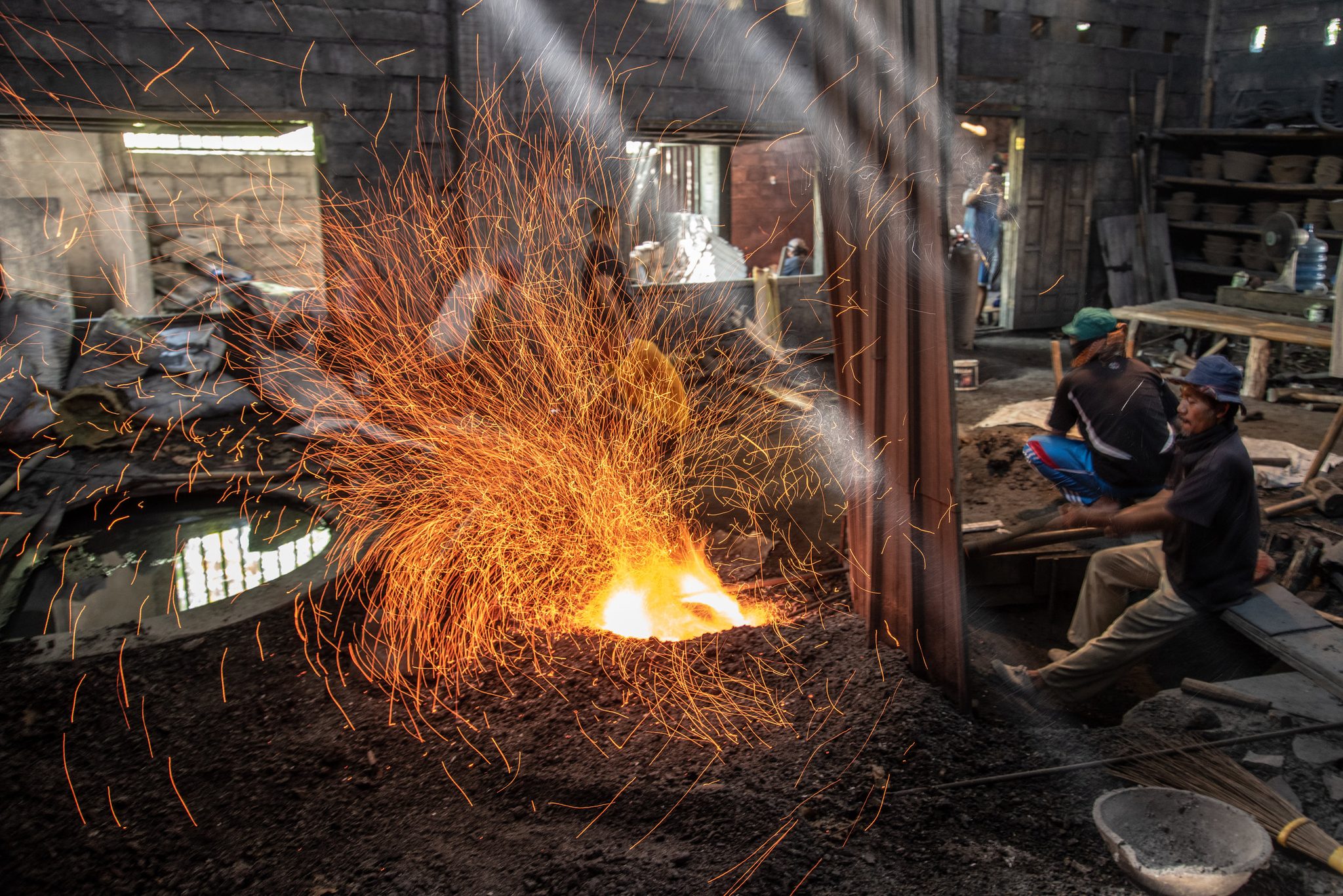 Sparks explode from the forge. Two workers wait out the heat on a bench.