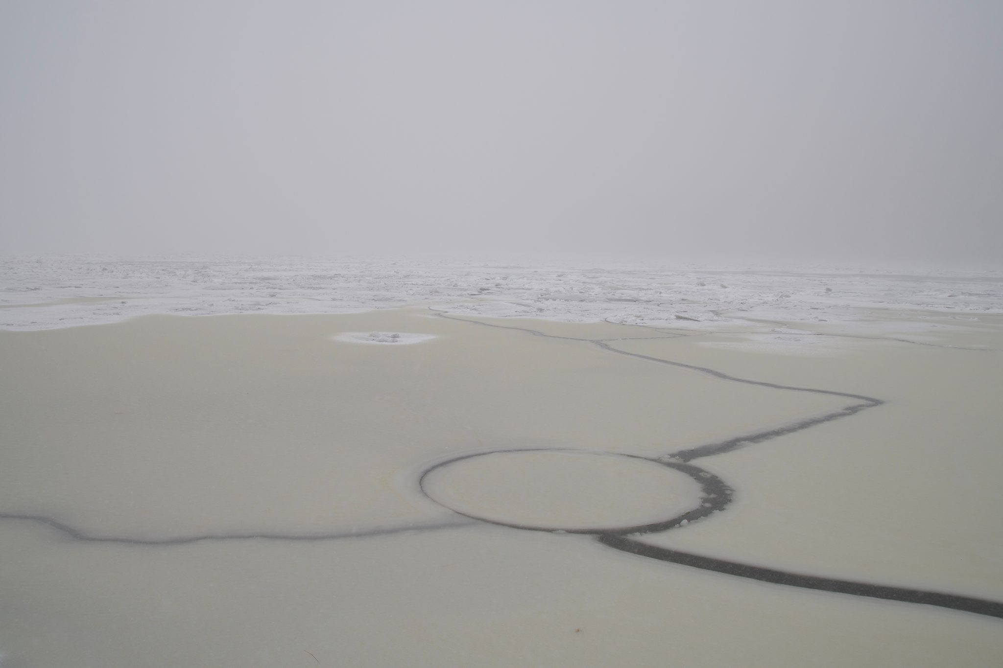 A ring crack drawn in the pale river ice under flat fog.