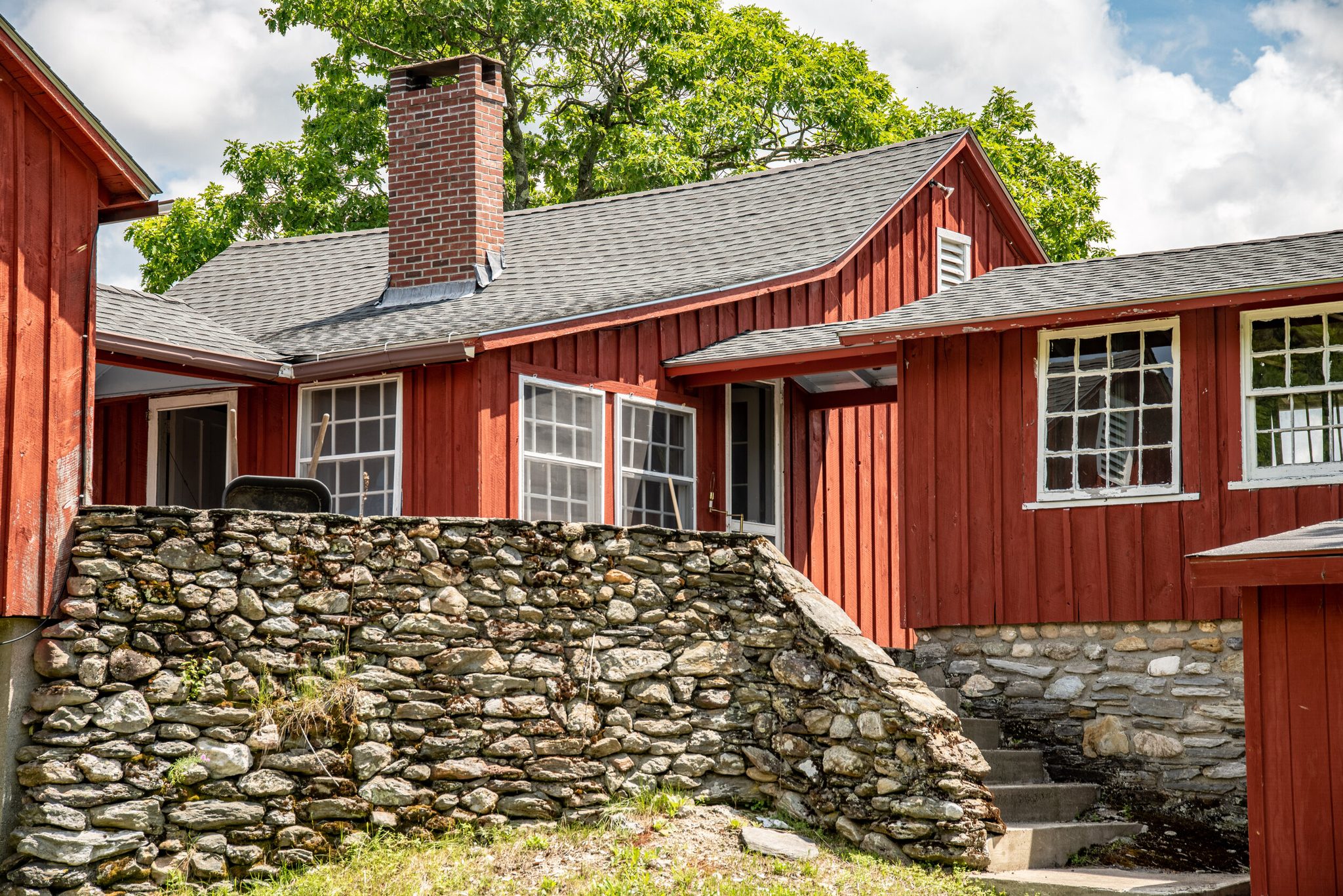 A red board-and-batten house rises from a fieldstone foundation, its chimney catching the sun.