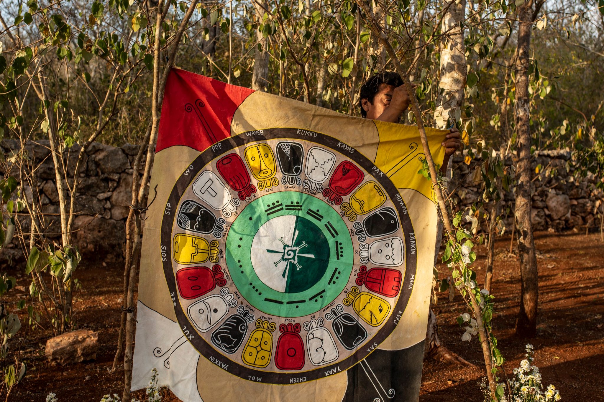 A man lifts a banner painted with the Maya calendar, its day signs circling a green heart.