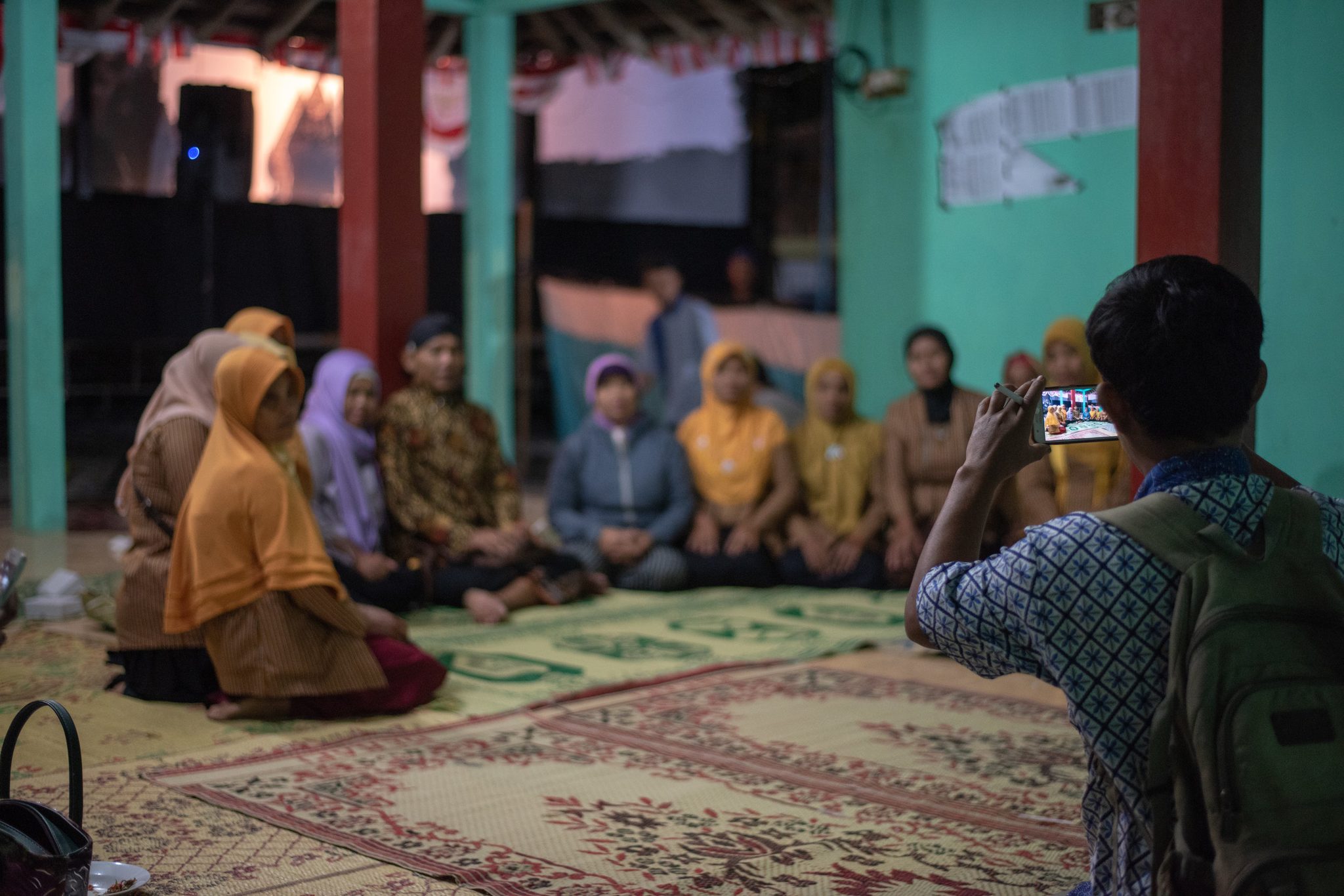 A man lifts his phone to photograph the women seated along the pavilion wall.