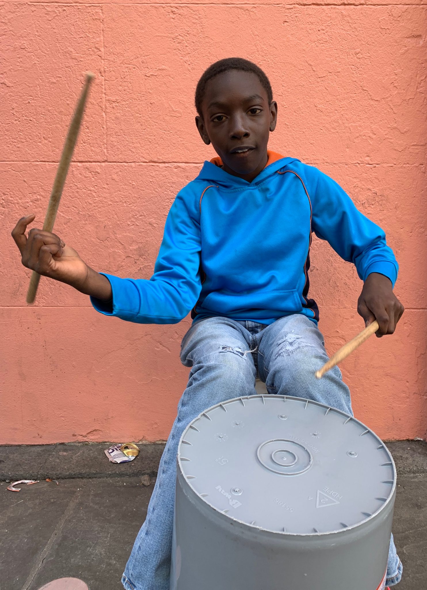 A boy in a blue hoodie drums on an upturned bucket against a salmon wall.
