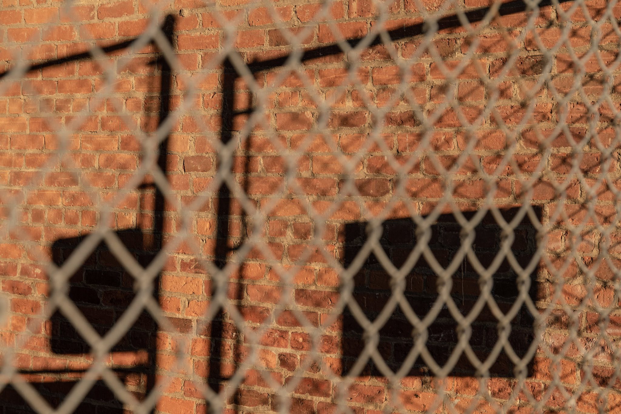 A chain-link fence casts its shadow on a sunlit brick wall.