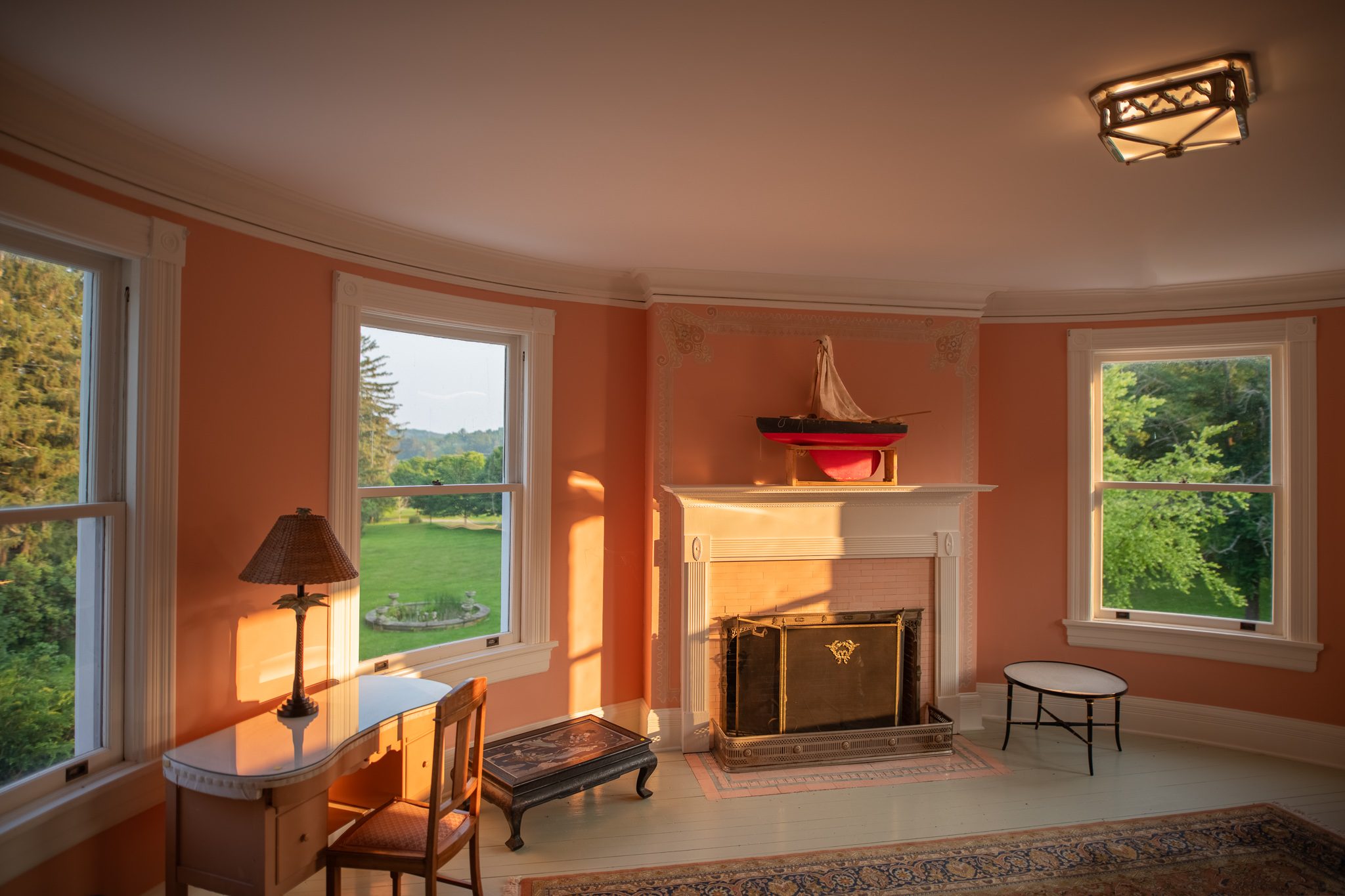 Low sun warms a salmon-colored corner room with a white mantel, a model boat above it and green lawn outside.