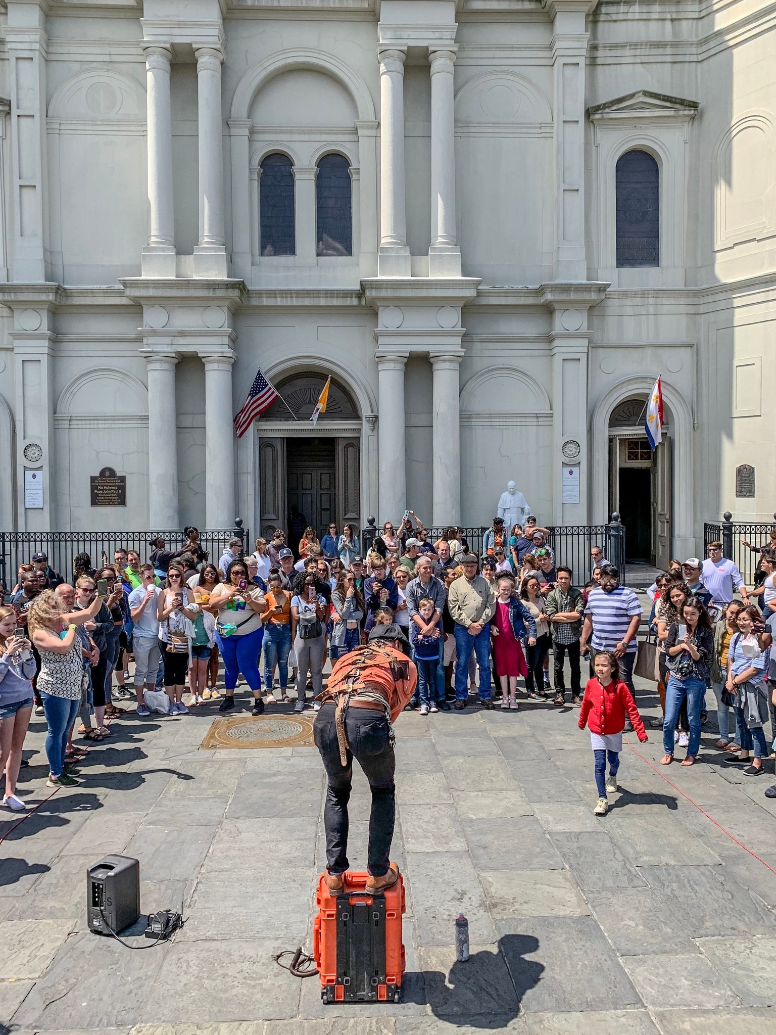 A street performer folds himself over an orange amp as the crowd leans in before the cathedral, and one small girl in red can't wait for the trick.