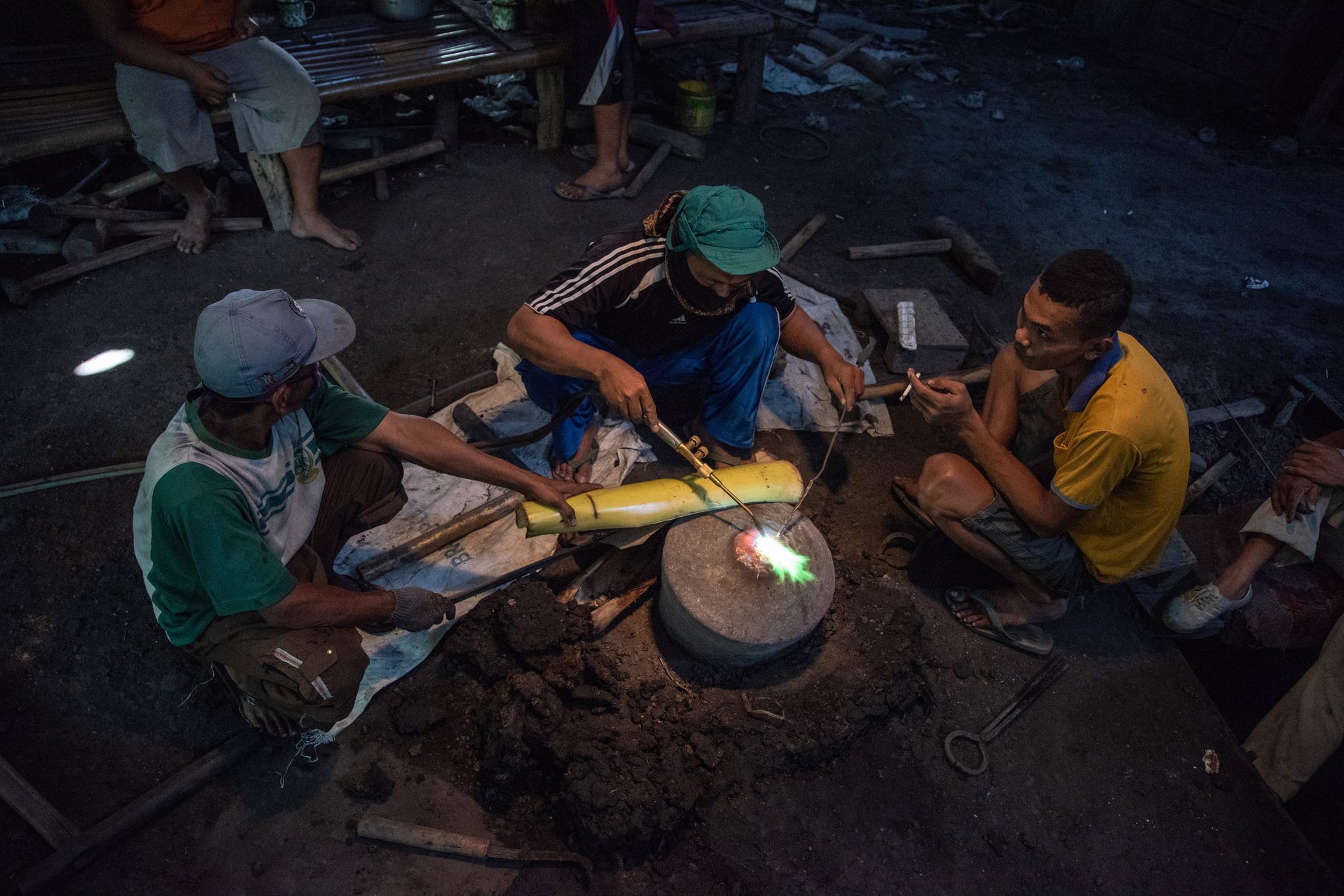 Three men crouch around the anvil as one of them works a new bar with the torch.