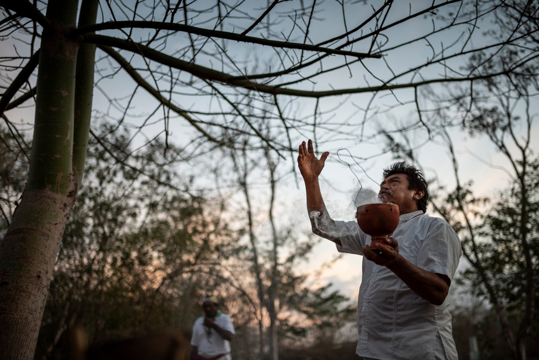 A man in white raises one hand to the sky and holds the offering bowl close with the other.