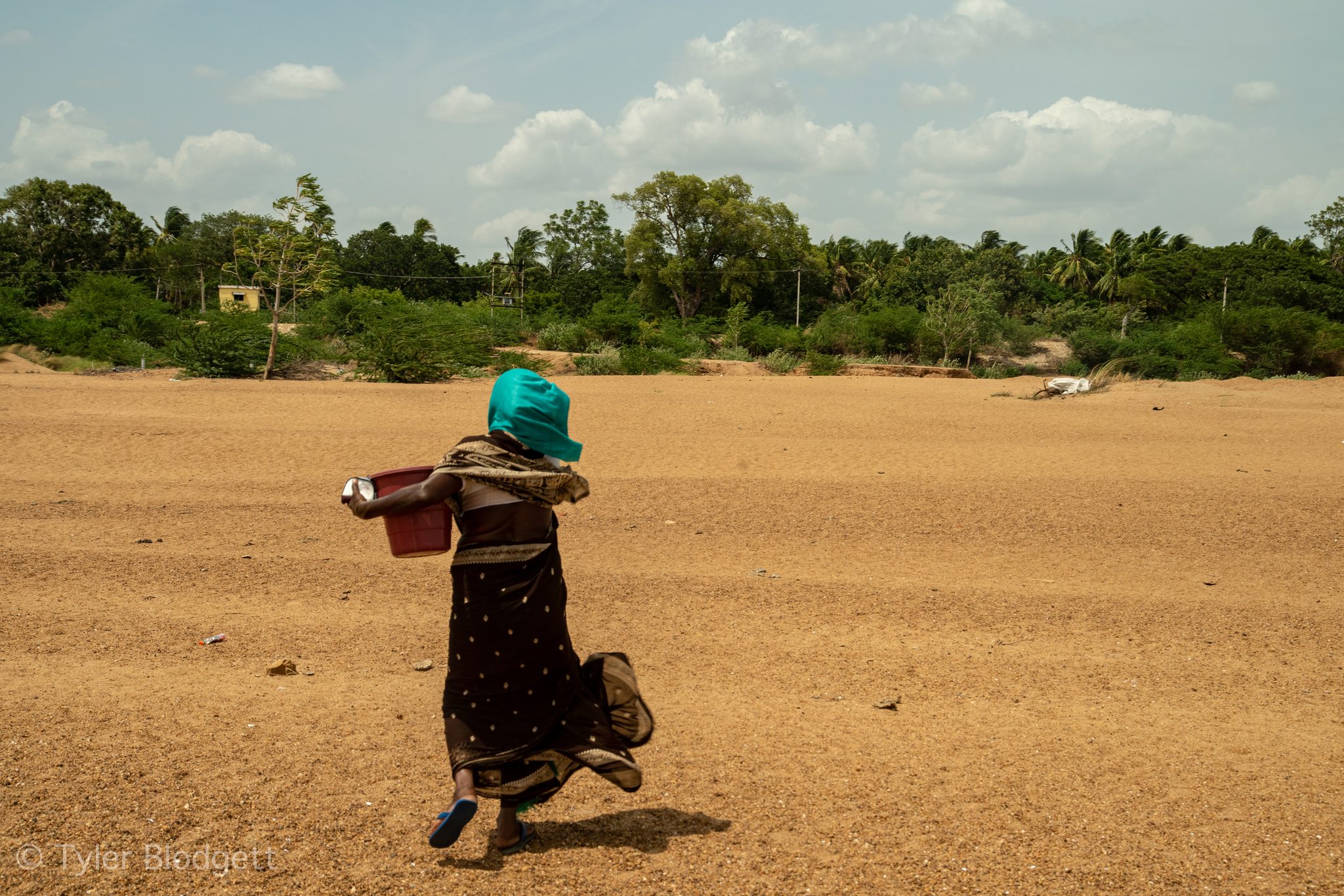 A woman in a teal headscarf strides across the dry riverbed with a red bucket.