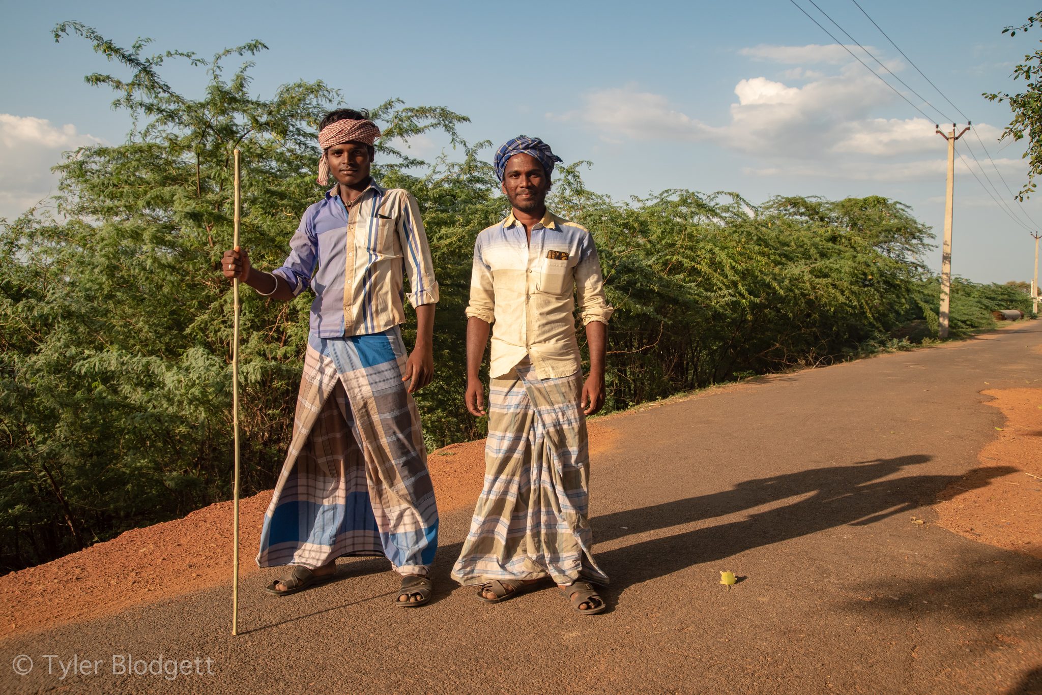 Two men in checked lungis stand on the red dirt road, one leaning on a staff.