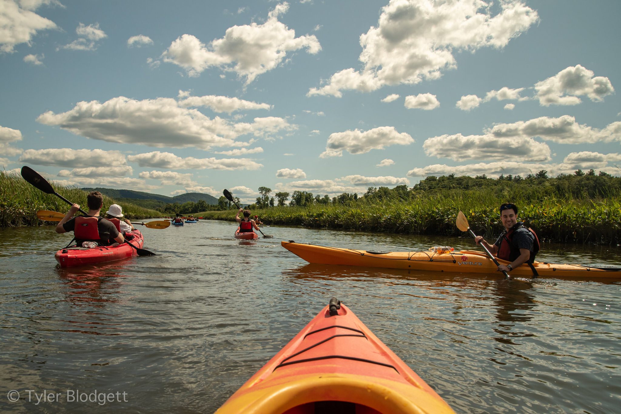 From the bow of a kayak, the group paddles up the marsh under summer clouds.