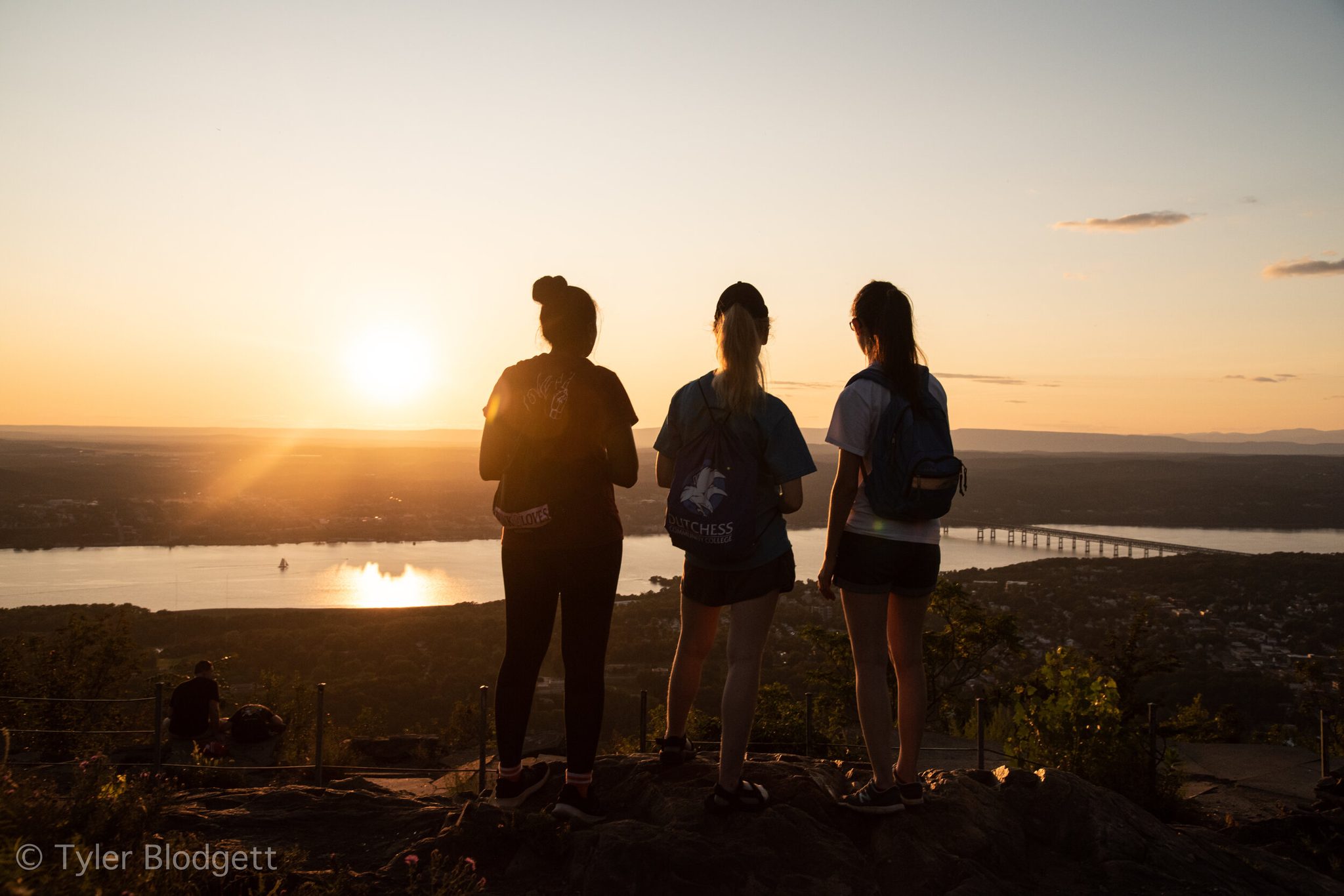 Three hikers stand on the ledge as the sun drops toward the river, the bridge a thin line of light below.