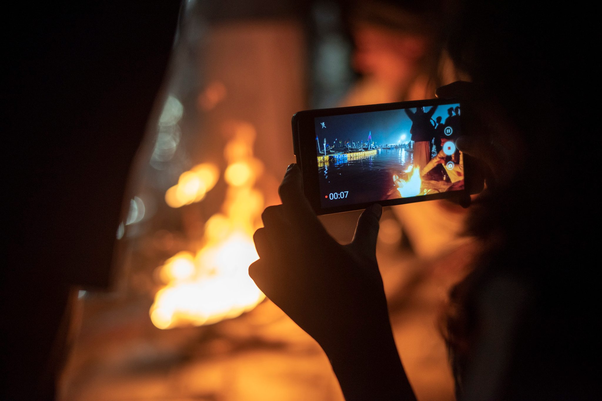 A phone records the fire and the lit shore beyond it.