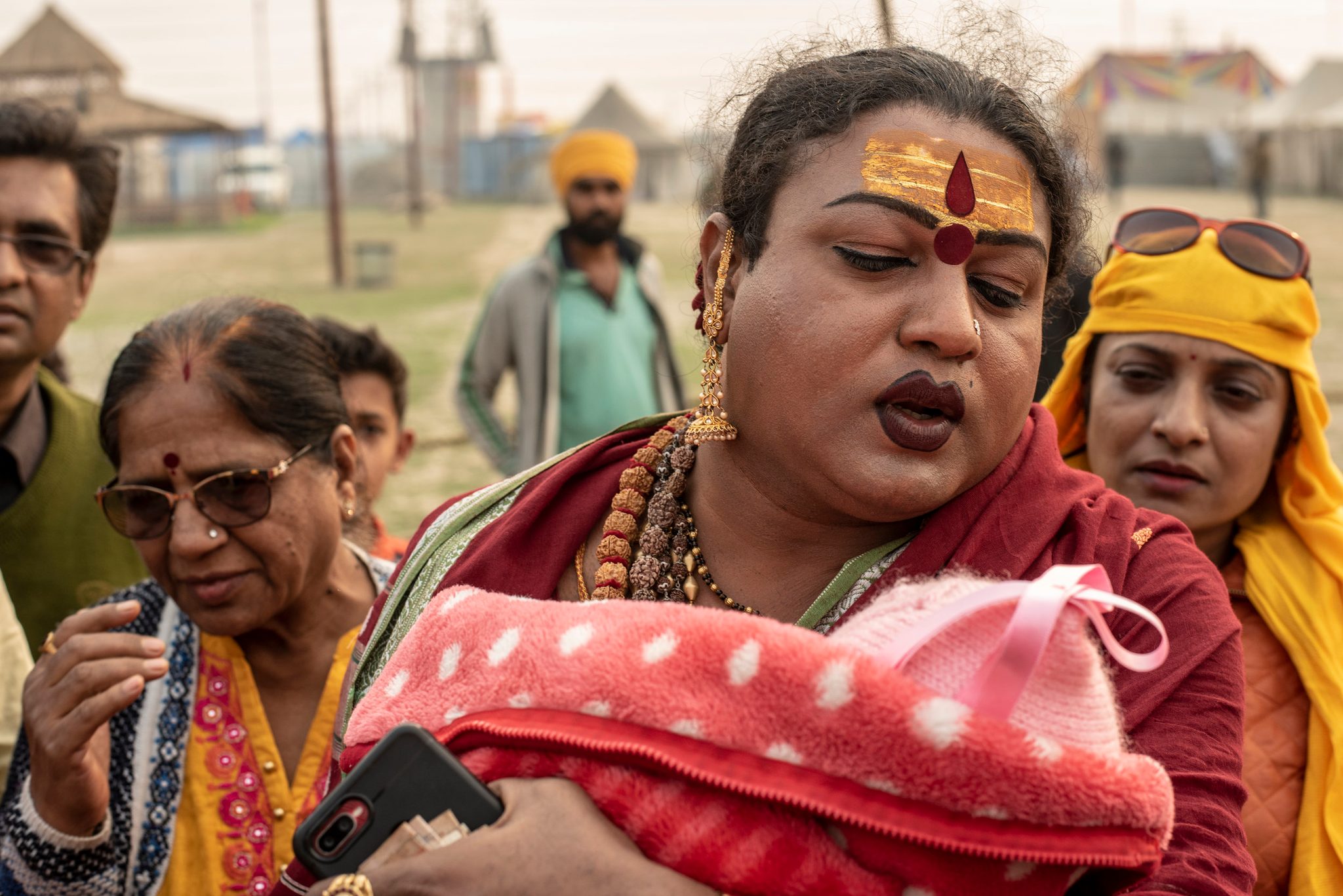Mayuri Ma cradles a baby in a pink blanket, her forehead painted gold and red, as the others lean in close.