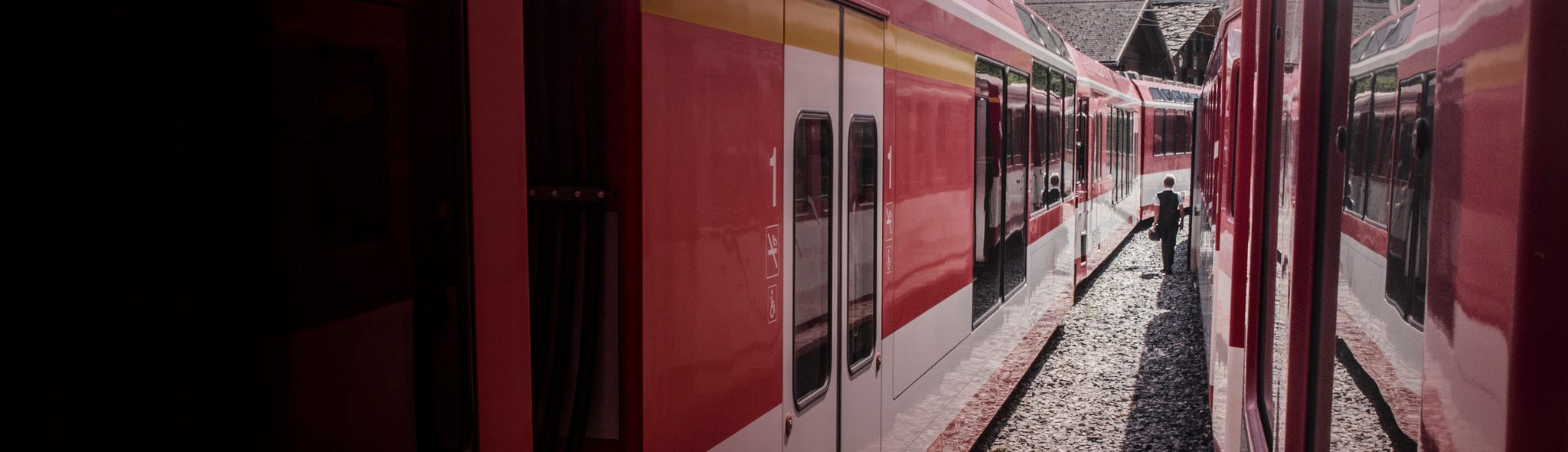 Between two red trains, a lone figure walks the gravel at the station.