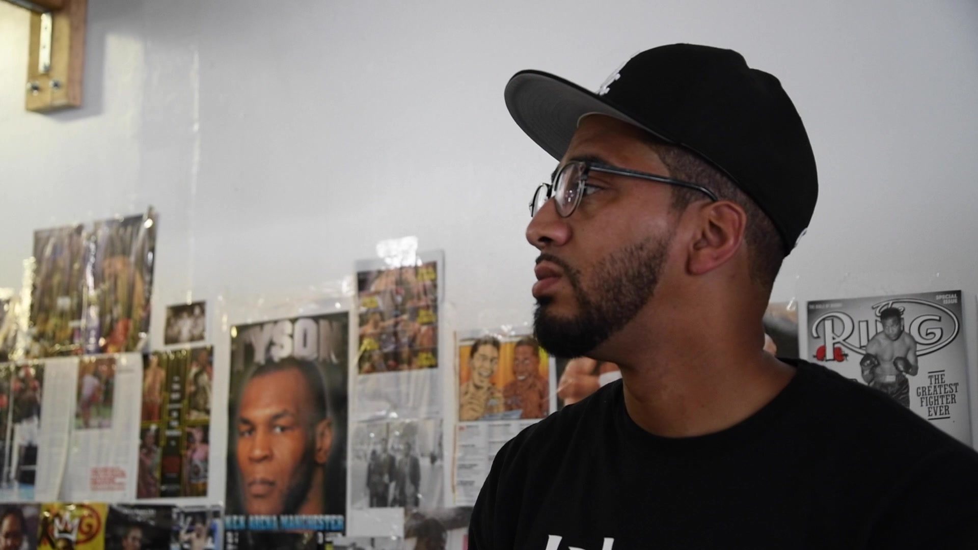 Against the poster wall, a man in a black cap watches the ring, a Tyson cover over his shoulder.