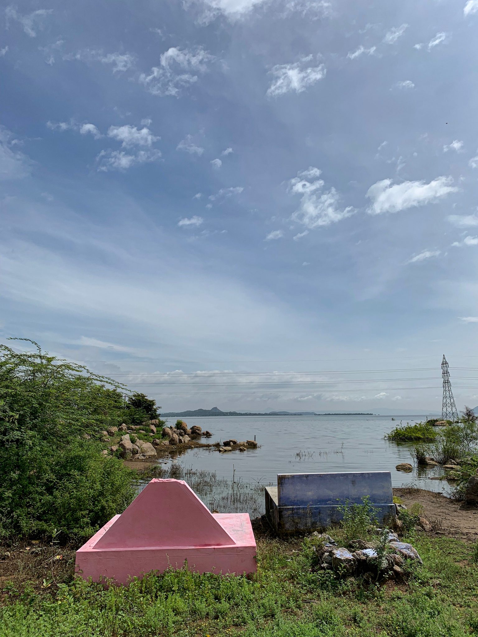Colorful burial pyres on the edge of the Kaveri, the water and a power pylon beyond.
