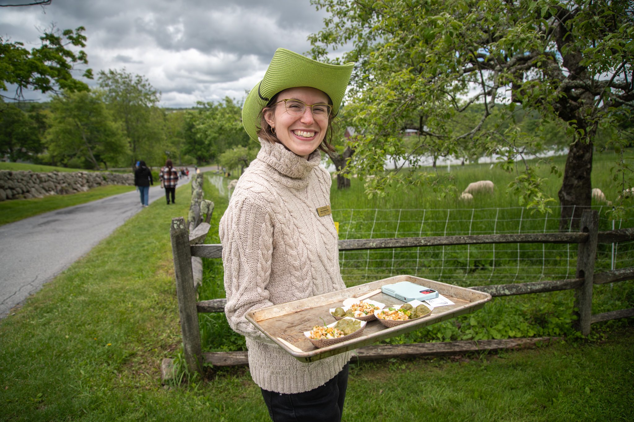 A volunteer in a green hat offers a tray of snacks by the pasture fence.