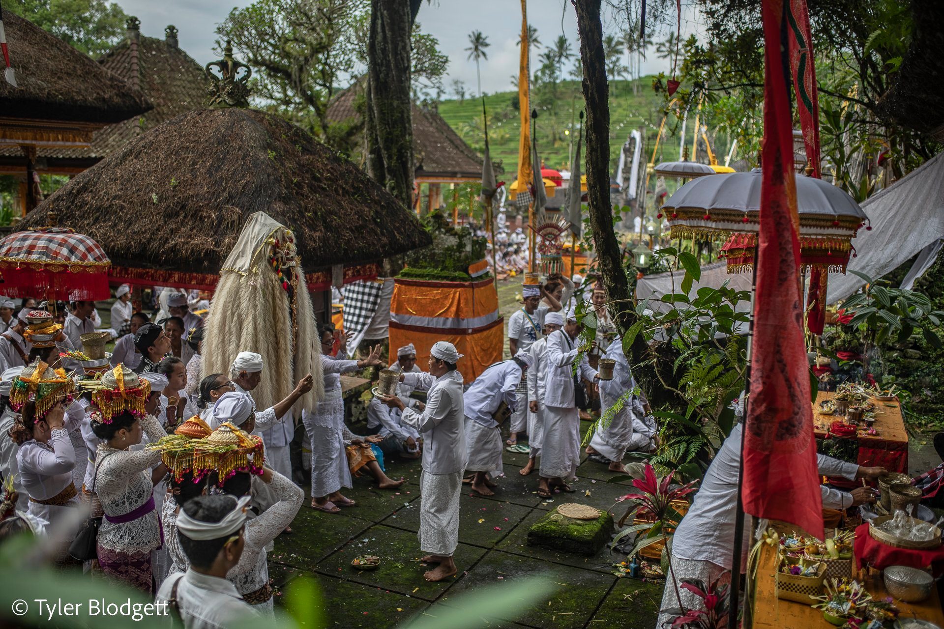 The procession winds between the thatched shrines, red banners hanging still in the gray light.