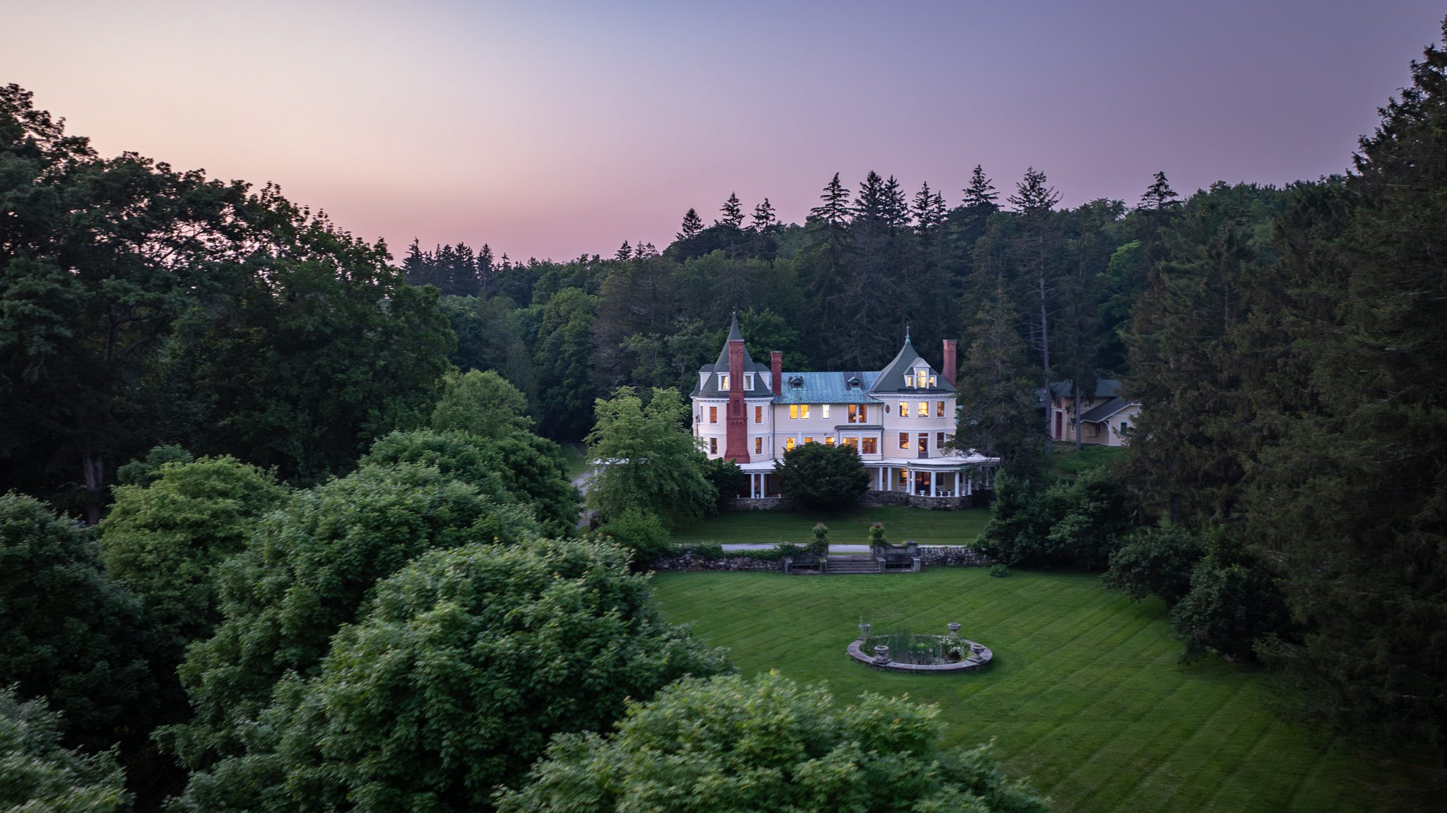 Dusk settles over the main house, every window lit, the lawn falling away toward the tree line.