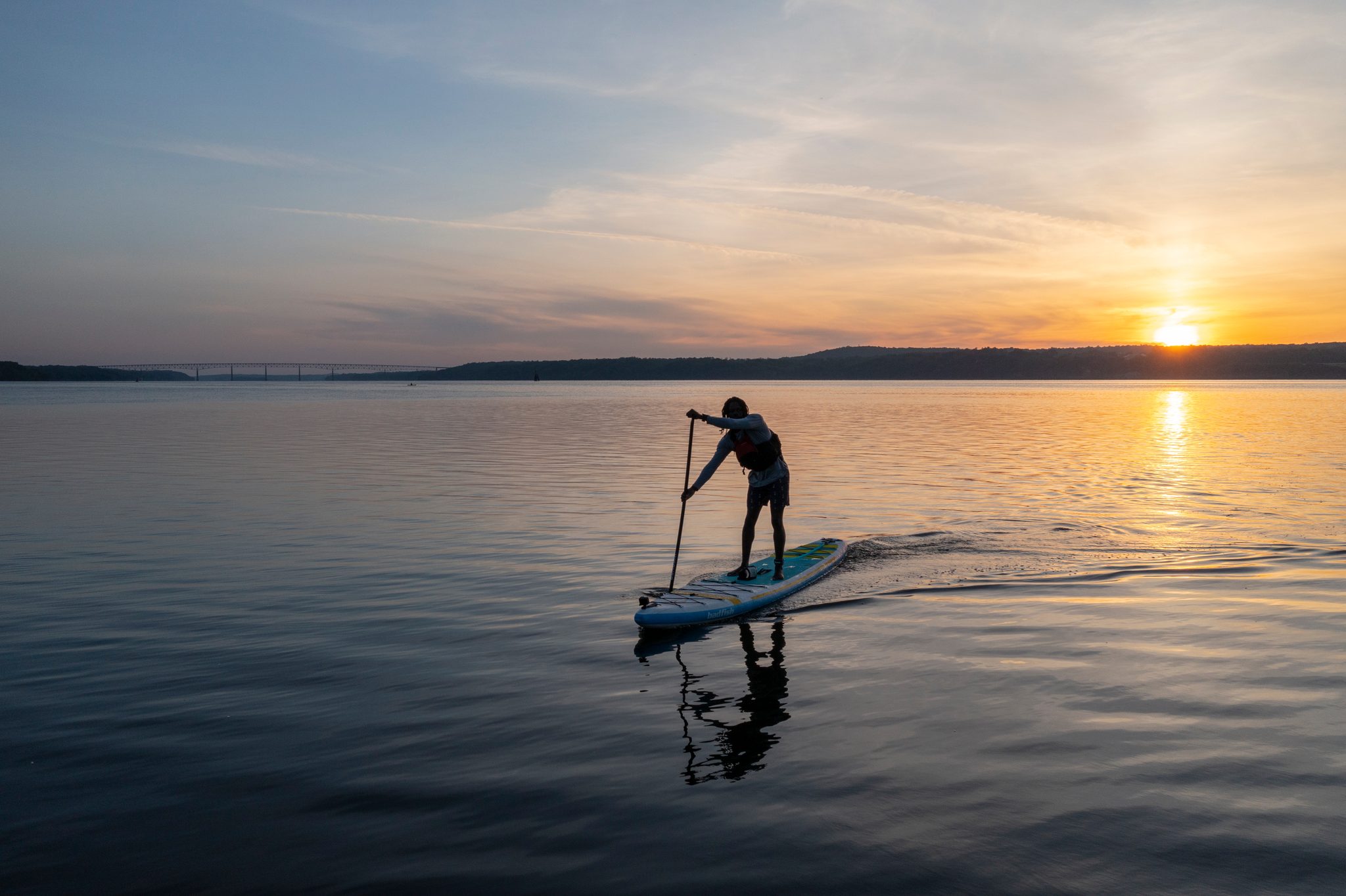 Standing tall on the board, a lone paddler strokes across mirror water with the sun low behind.