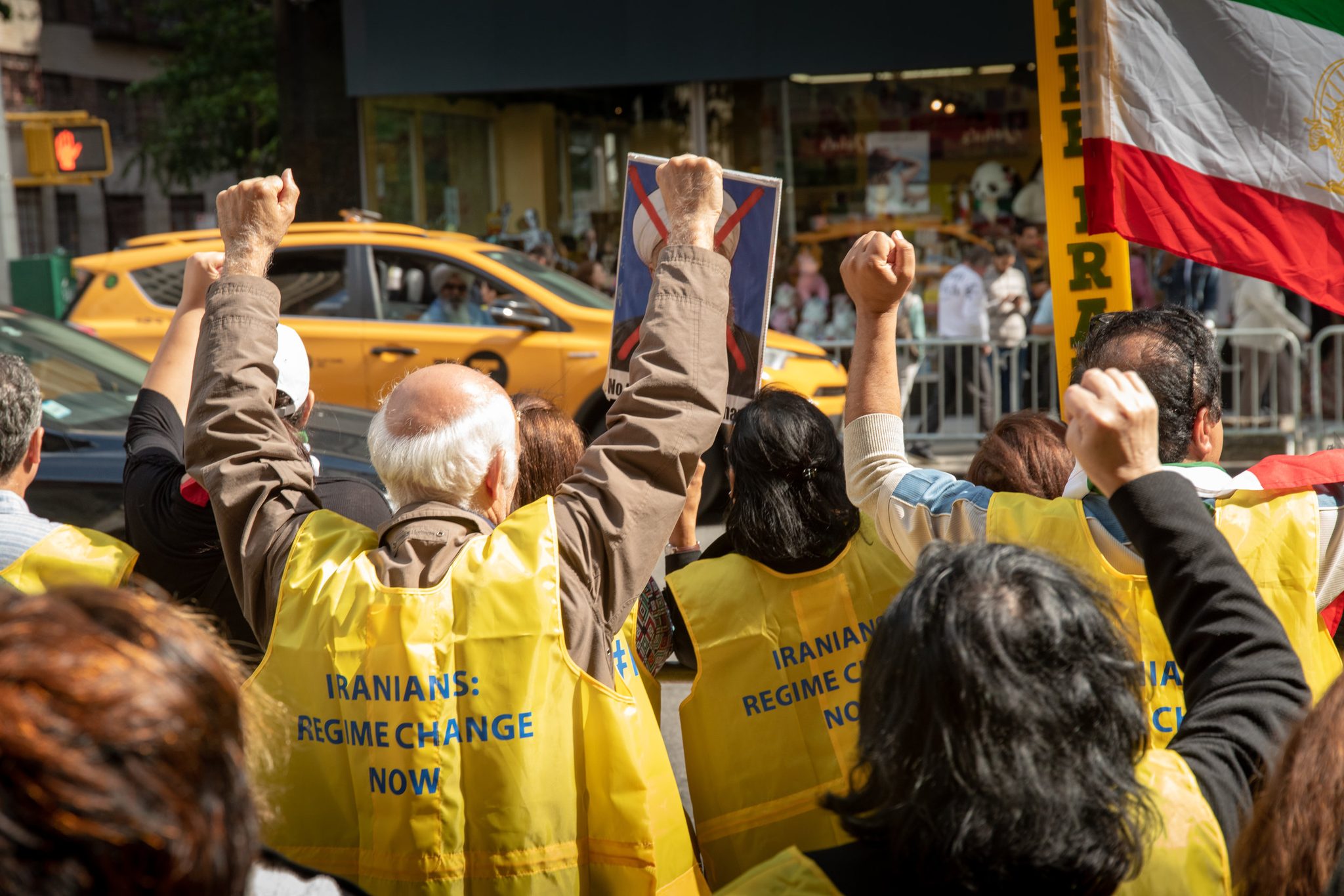Fists and placards rise over yellow vests reading Iranians: Regime Change Now, the old lion-and-sun flag above them.