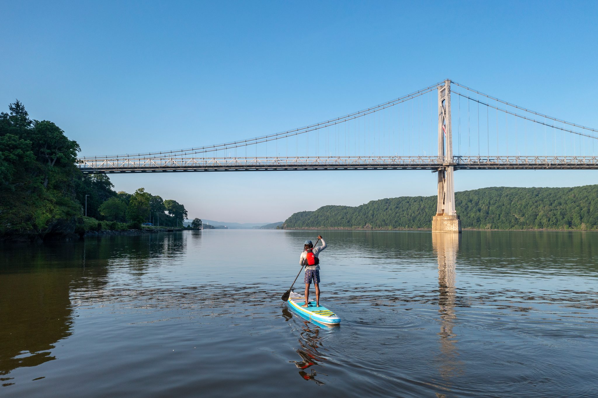The board glides across calm blue water under a suspension bridge, the far shore still in shadow.