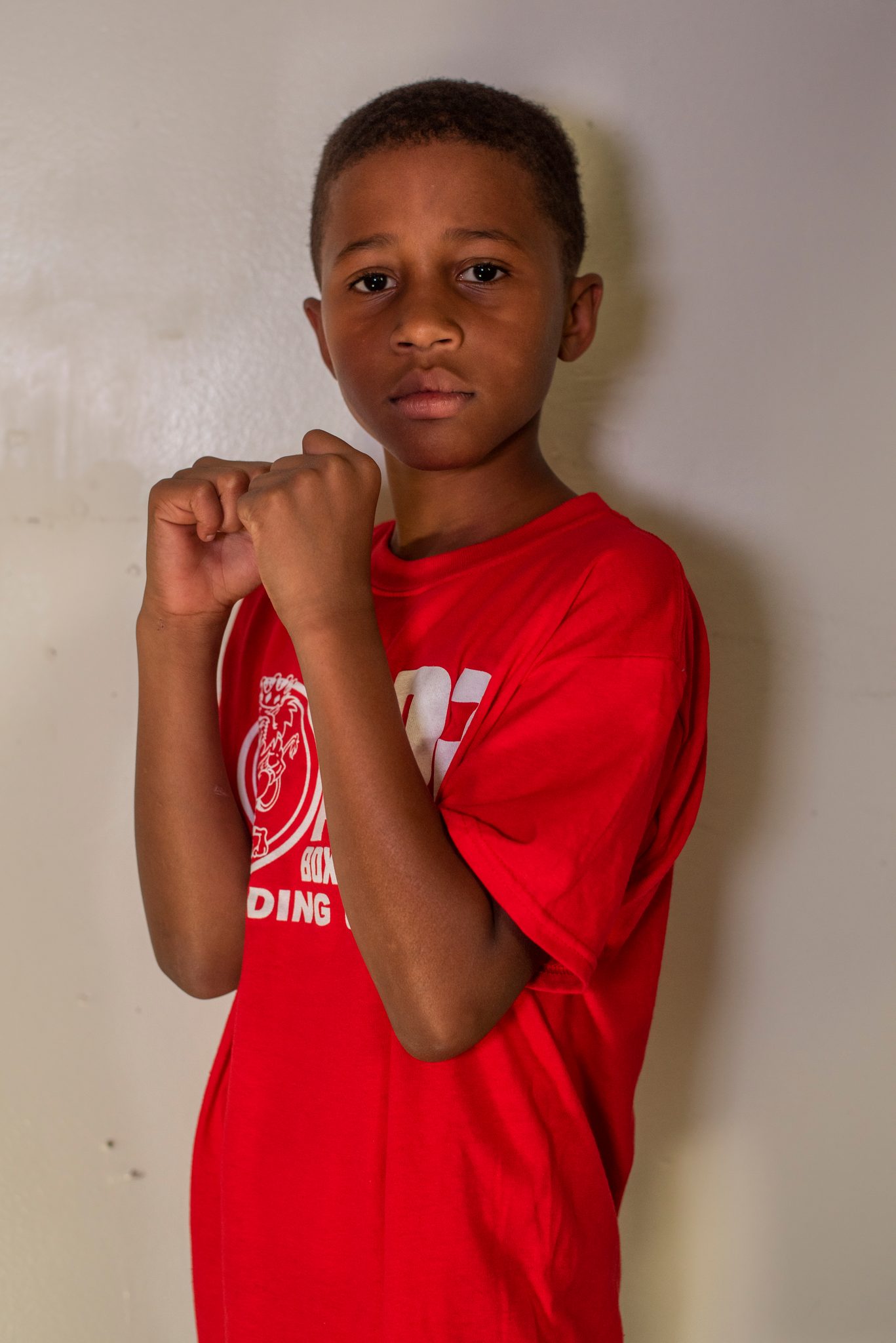 Against the white wall, a young boxer brings his fists up.