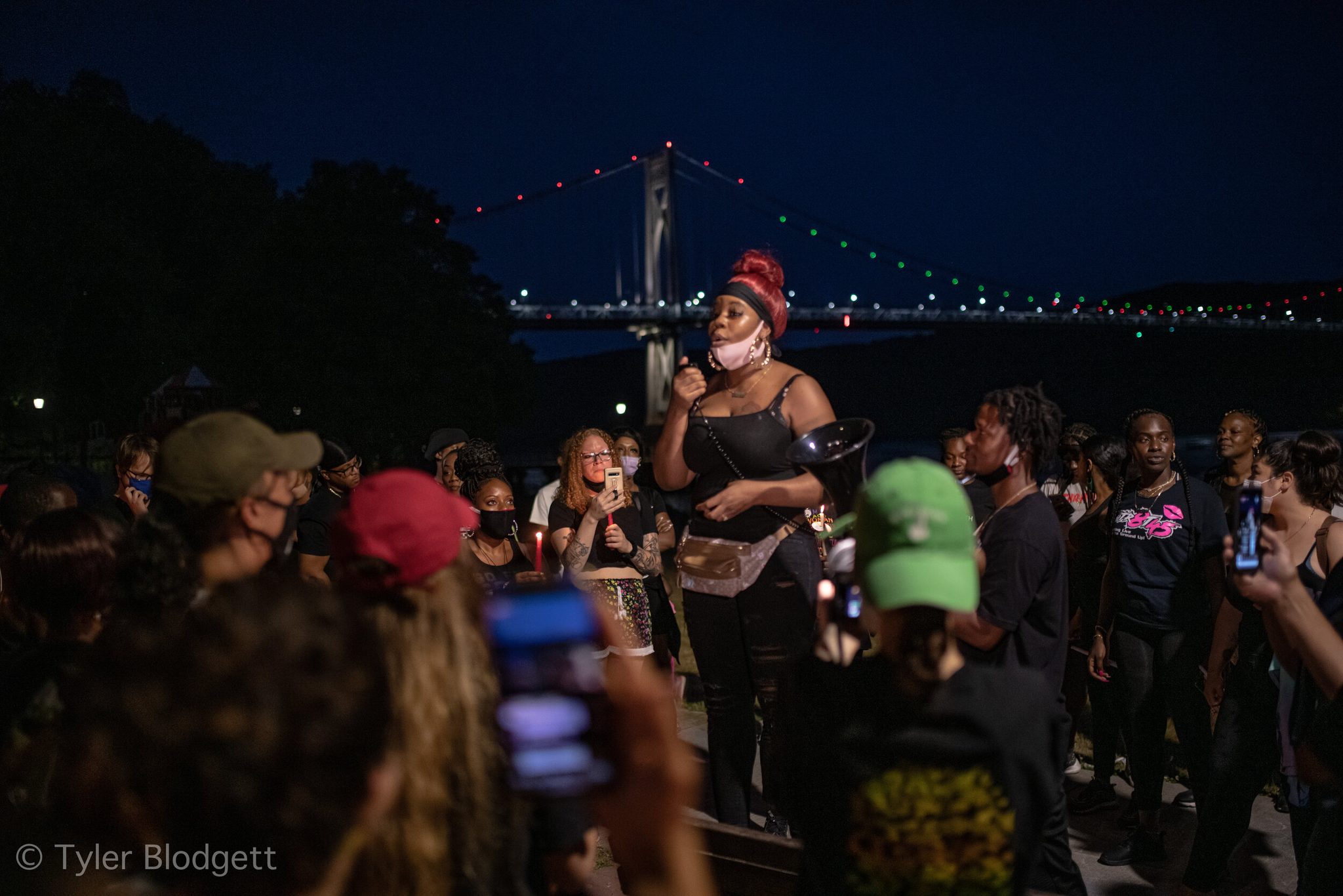 A woman speaks to the crowd by the river, the Mid-Hudson Bridge lit up behind her.