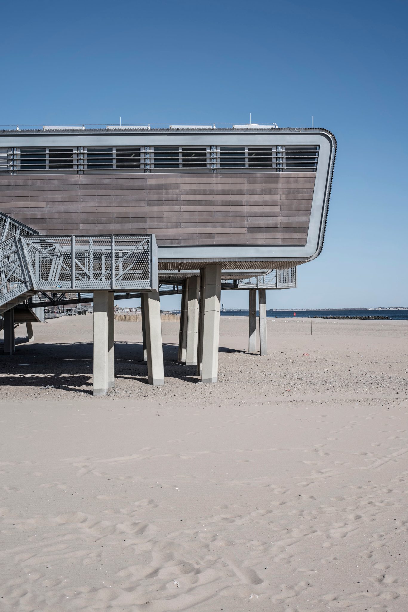 A pavilion on stilts rises from an empty beach under a clear sky.