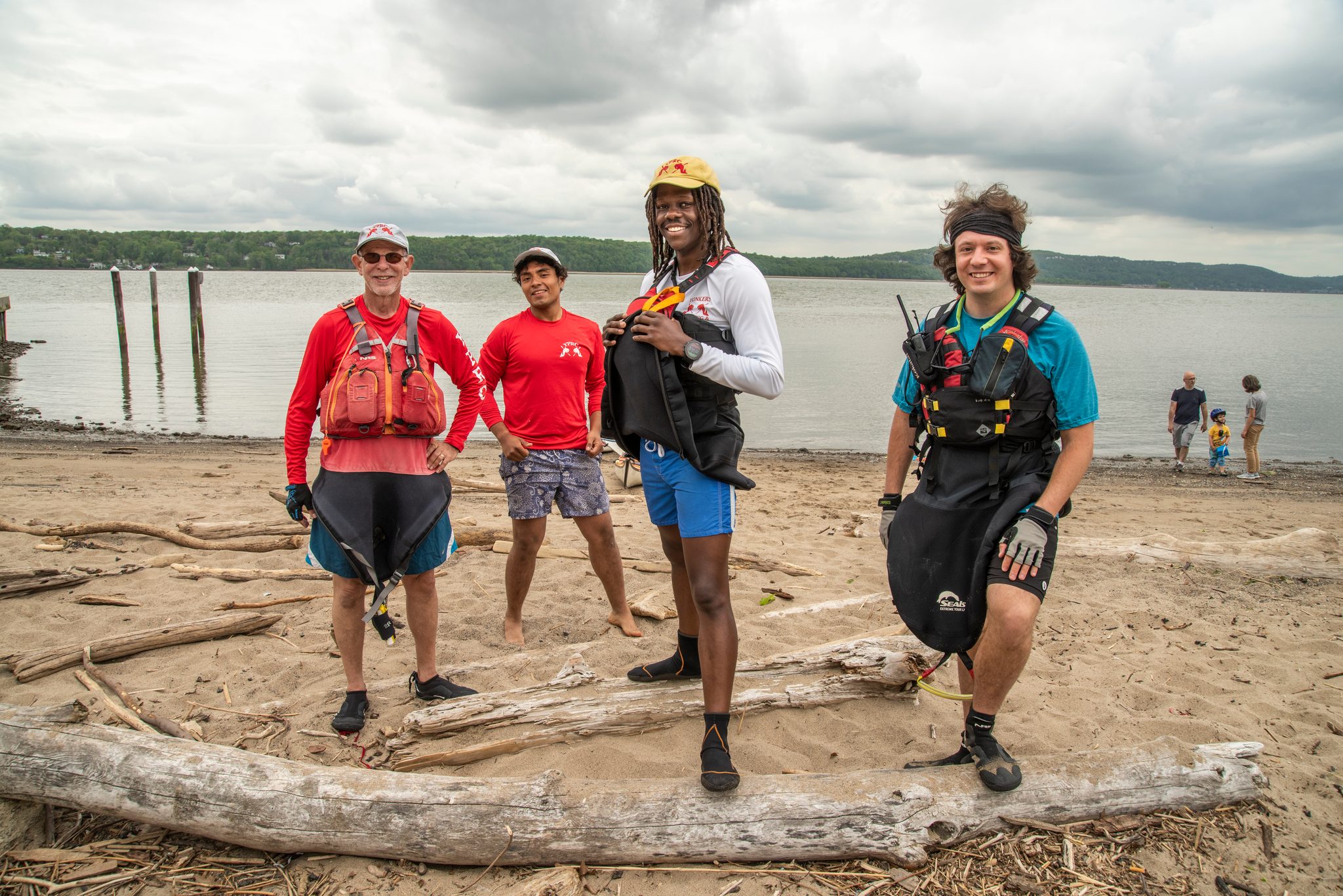 Four paddlers in life vests on a driftwood beach, Chev in the yellow cap at the center of the line.