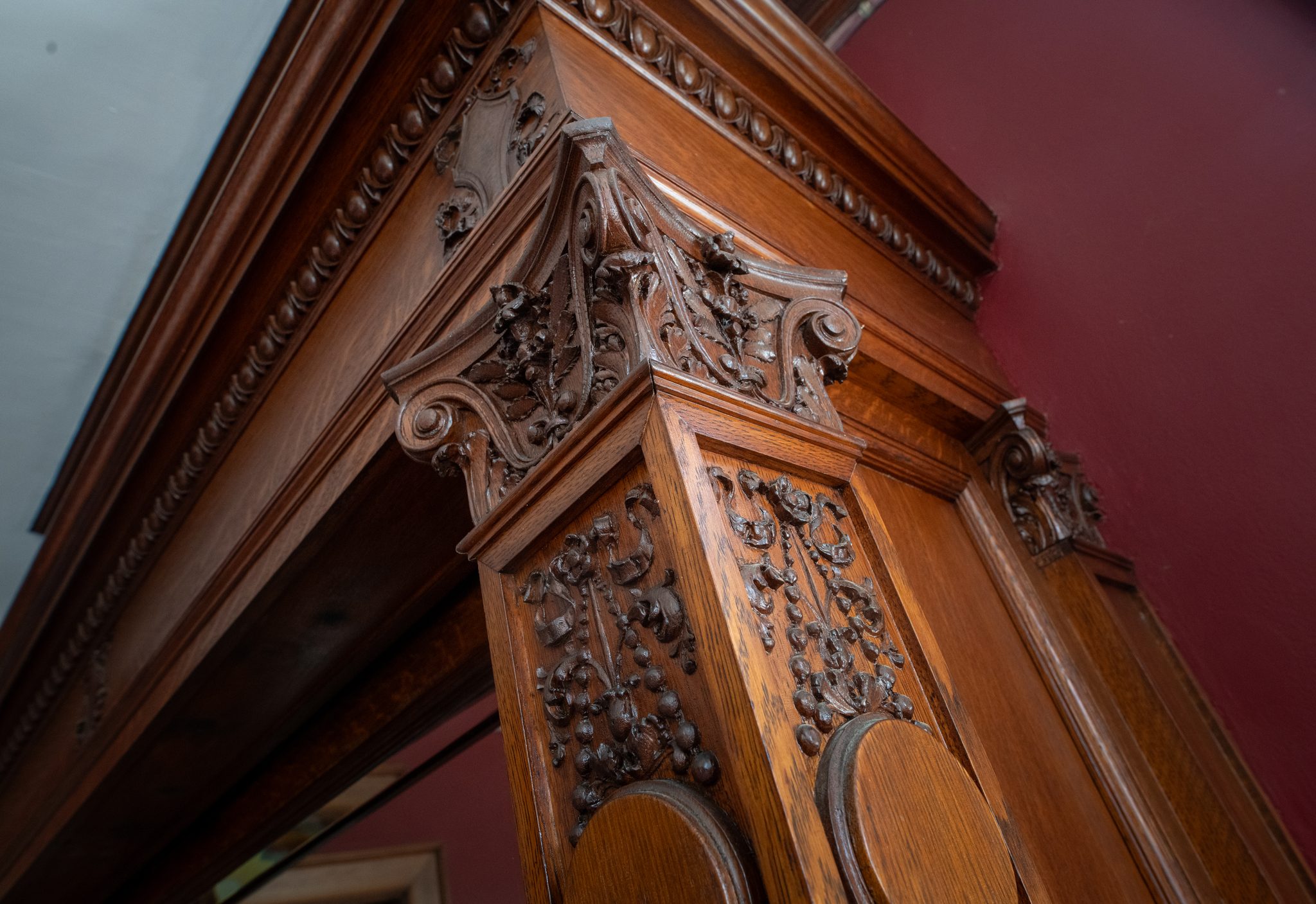 A carved oak pilaster against a red wall, the capital thick with leaves, scrolls and hanging clusters of fruit.