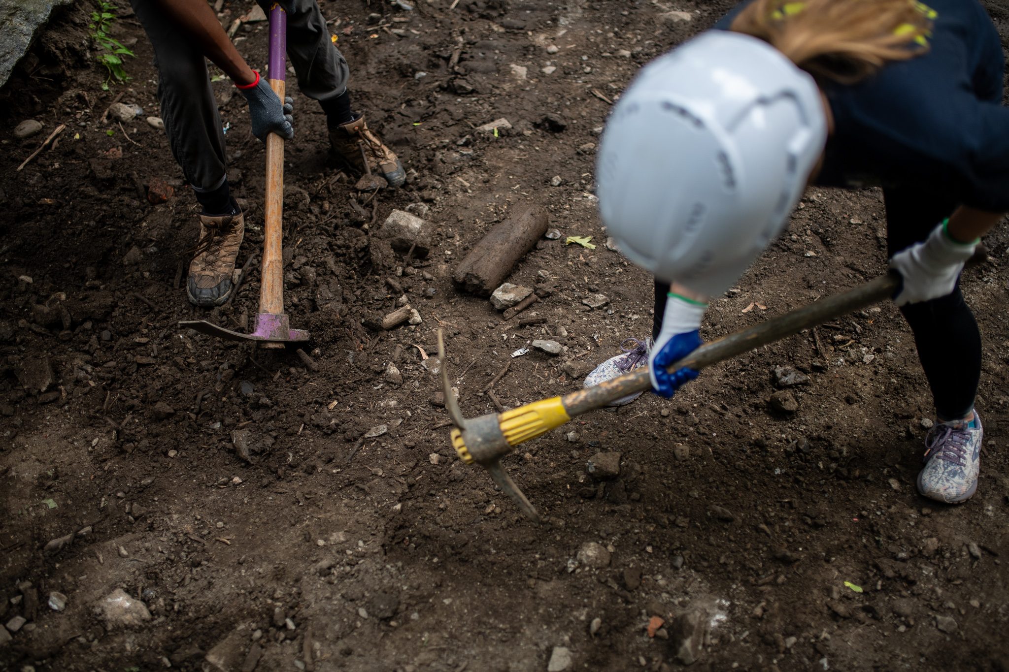 A pick swings into the dirt beside a white helmet.