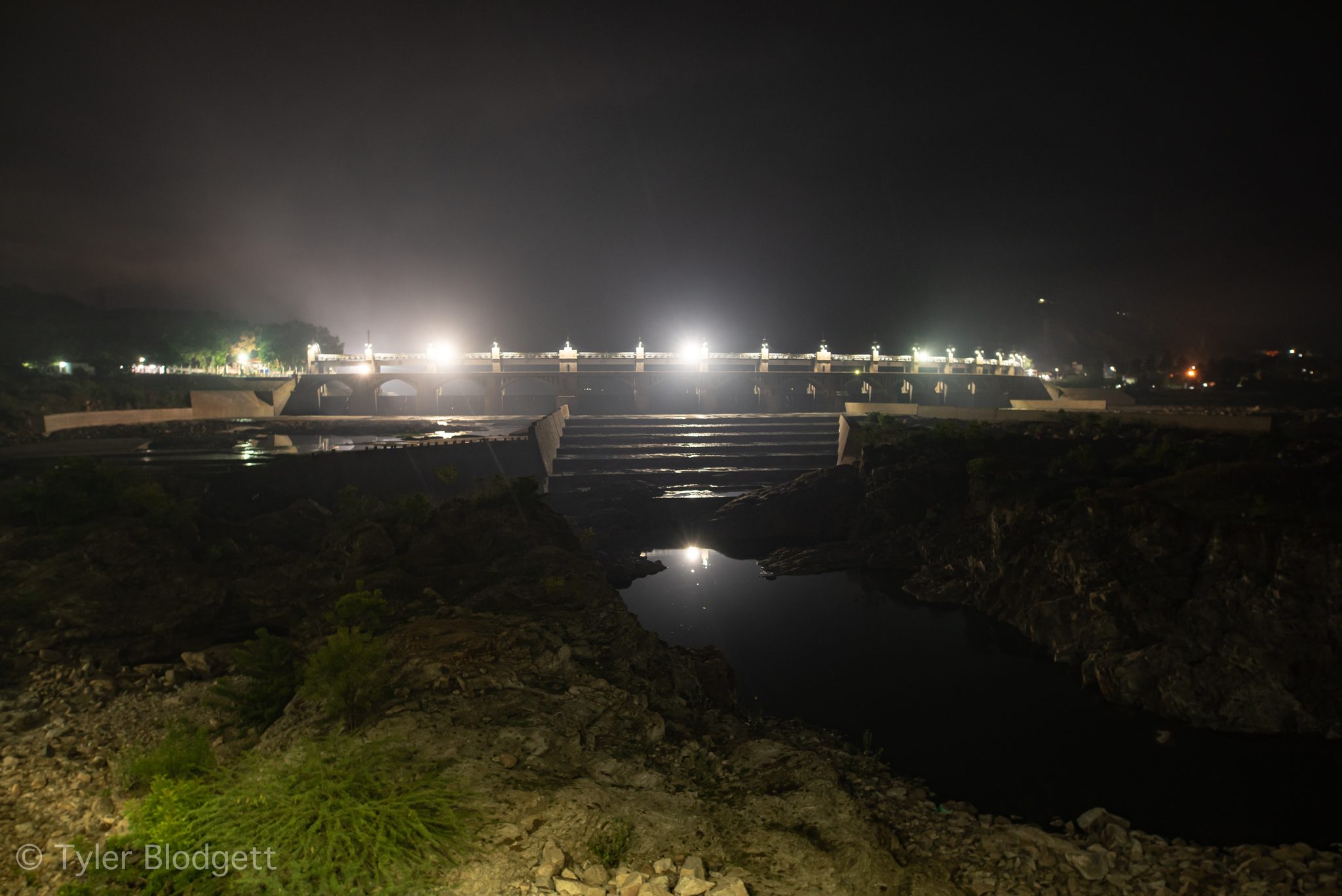 Floodlights blaze along the dam at night, the water black below.
