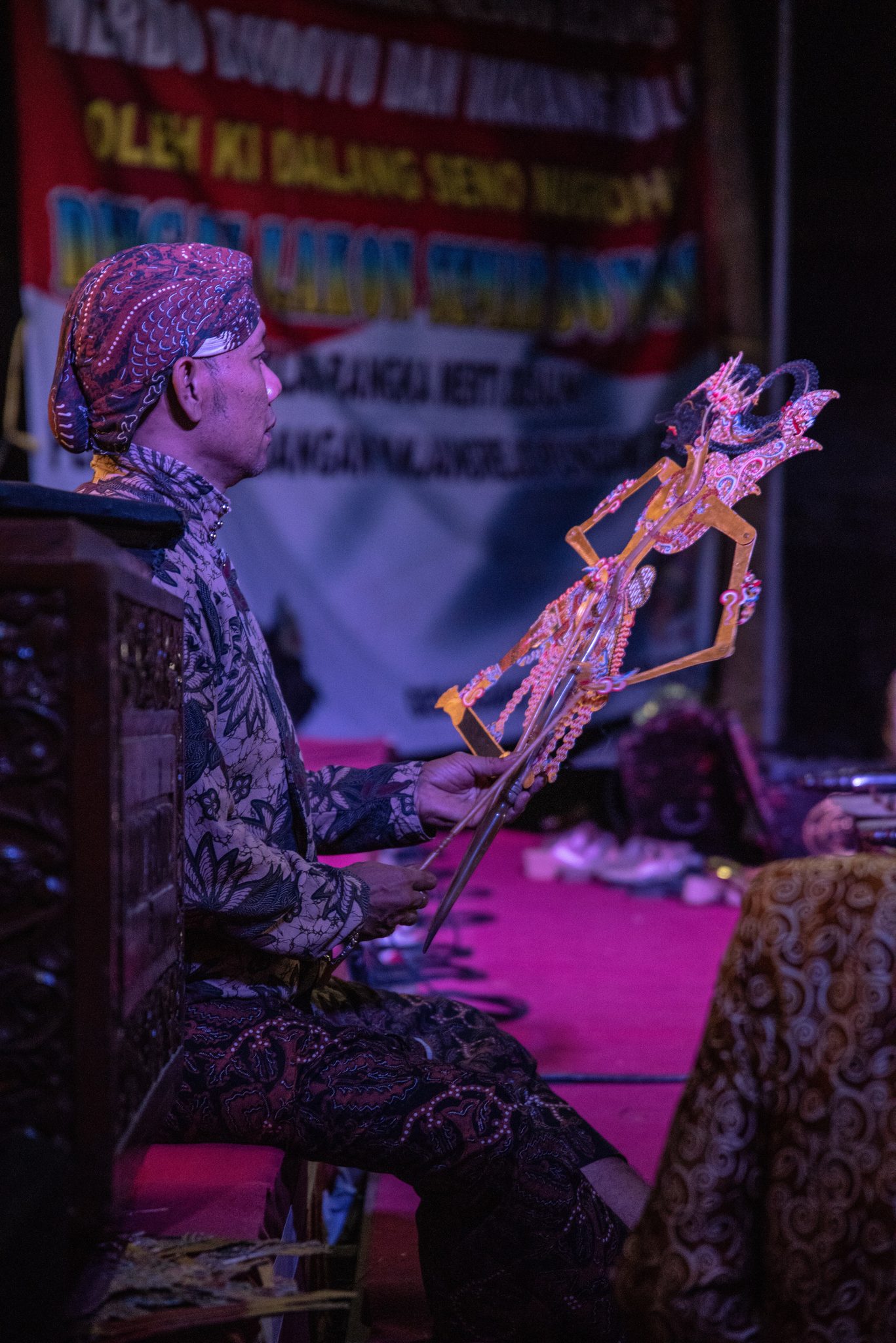 Ki Seno Nugroho lifts a puppet from beside the screen, beneath a banner bearing his name.