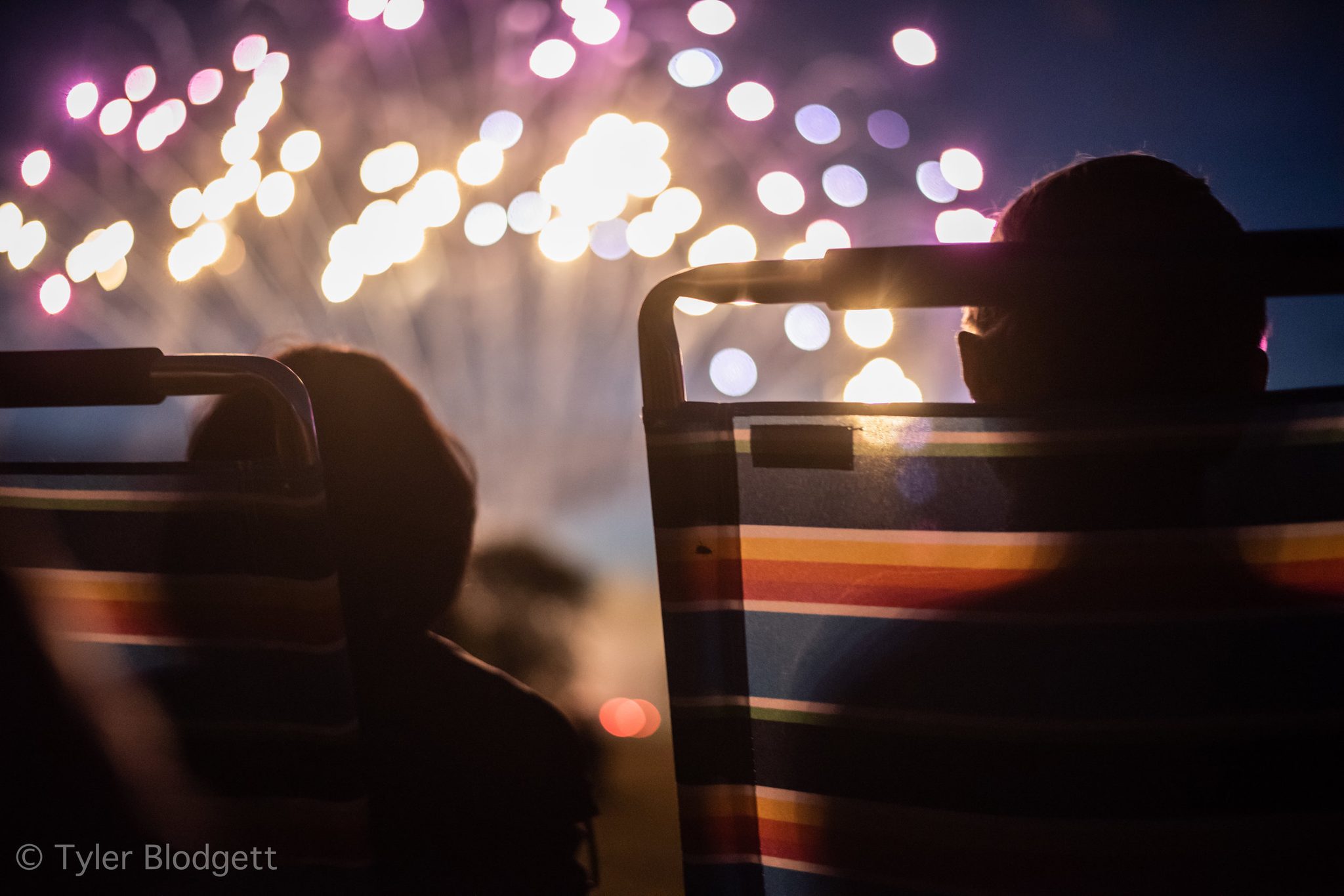 Heads tip back in folding chairs as the fireworks bloom out of focus overhead.