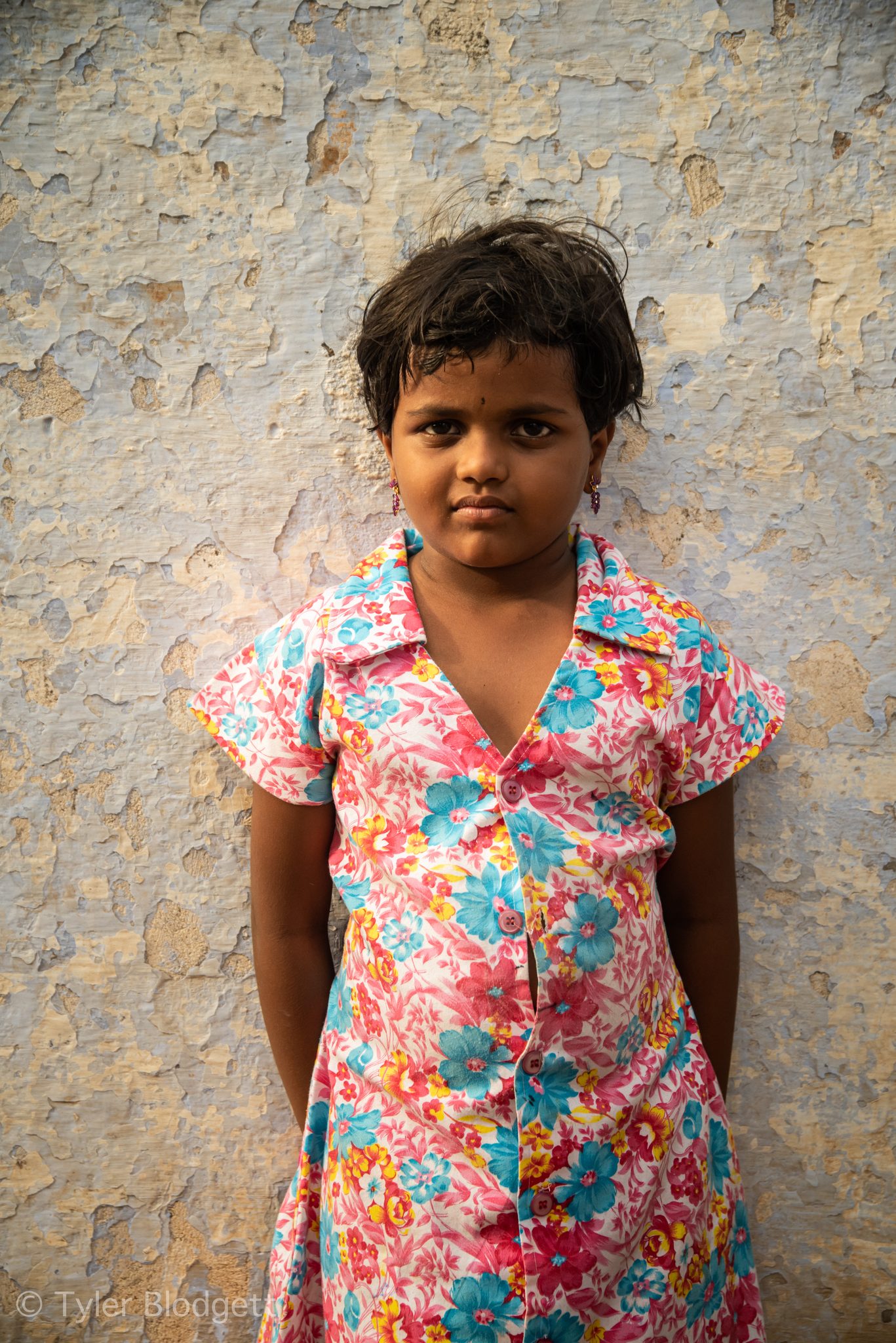 A girl in a flowered dress stands against a weathered wall.