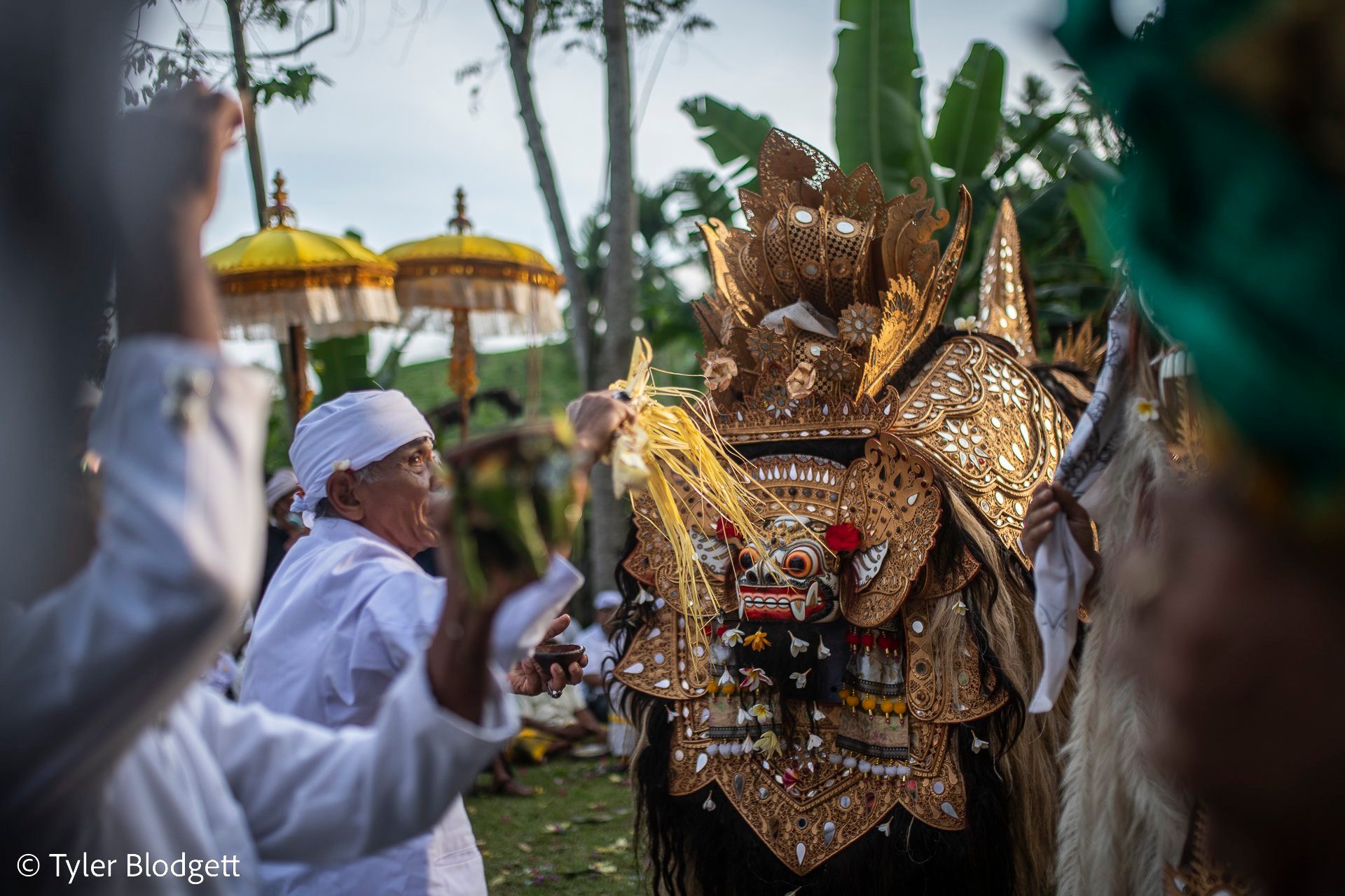 A gilded mask looms beside an old man in white, its carved teeth bared.