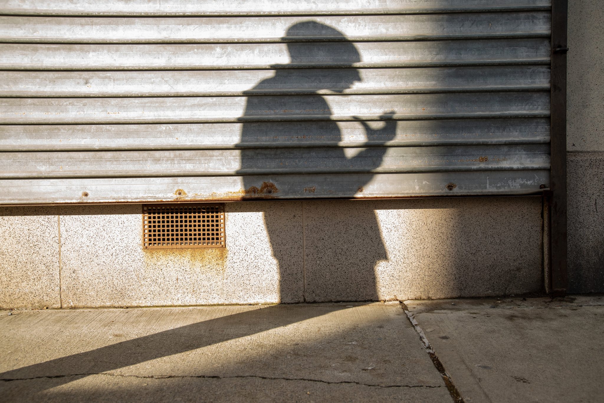 A passerby's shadow lifts a cup against a rolled steel shutter, the winter sun doing the rest.