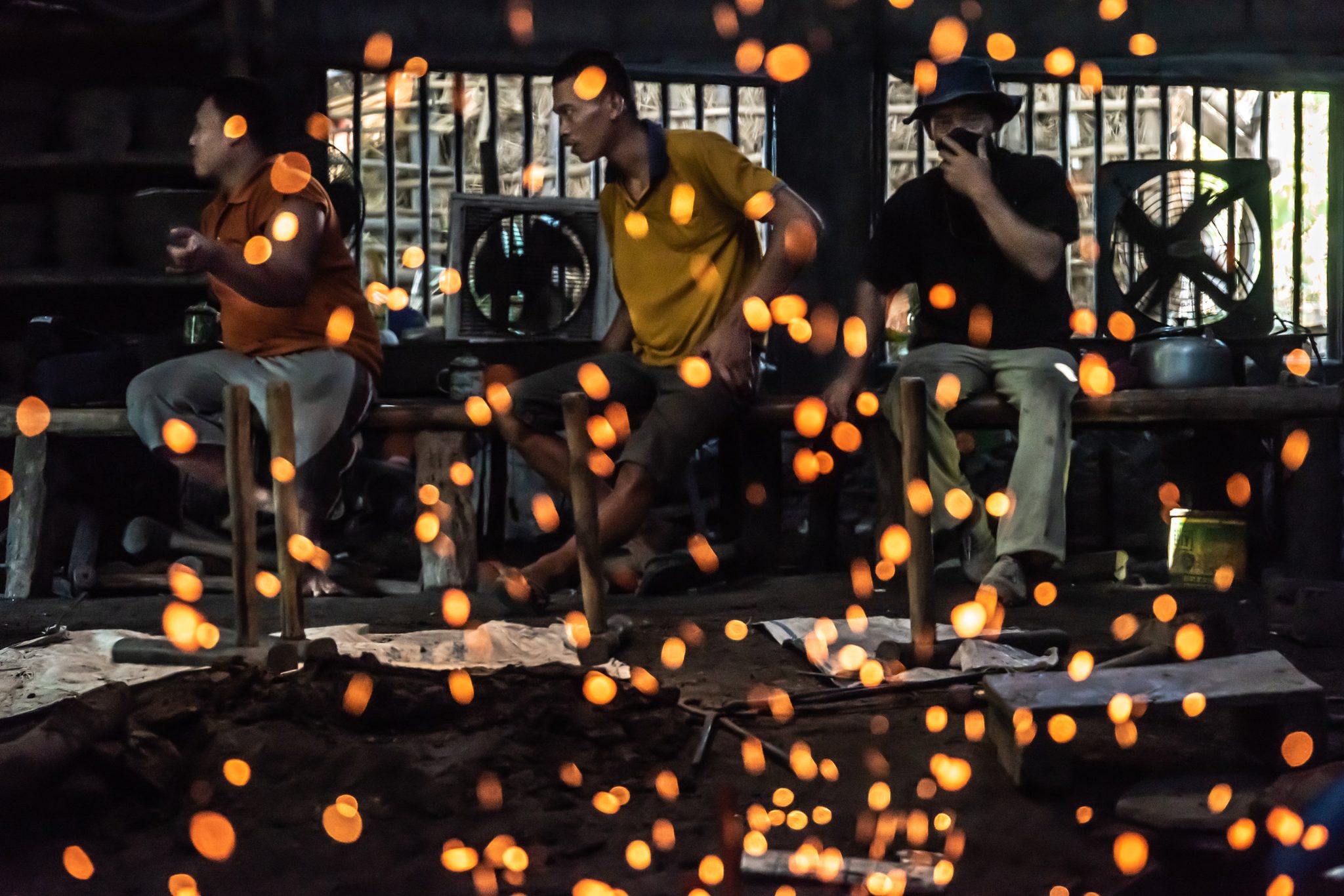Sparks drift like fireflies past the blacksmiths waiting on their bench.