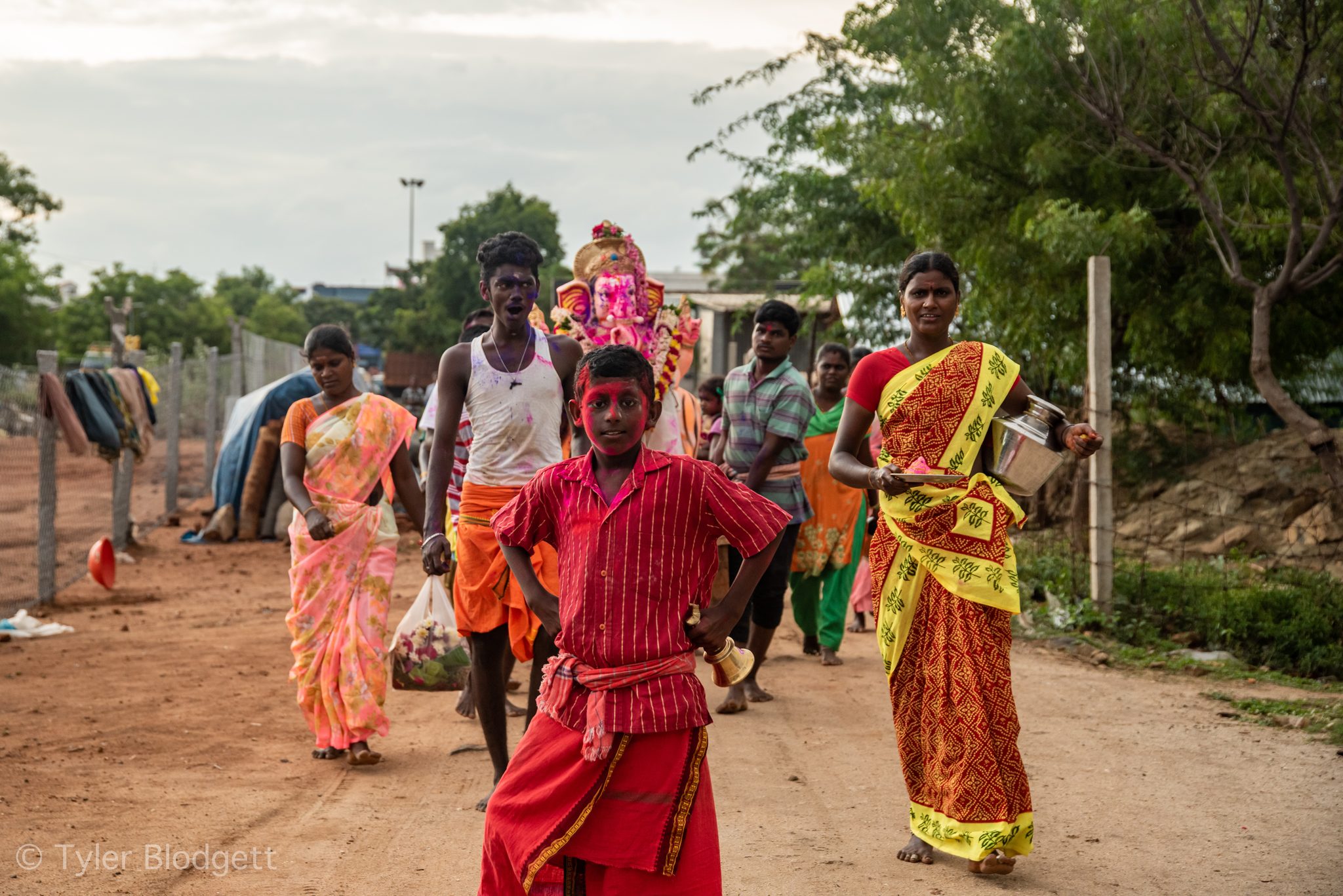 The procession comes down the dirt road, a boy in red leading the way.