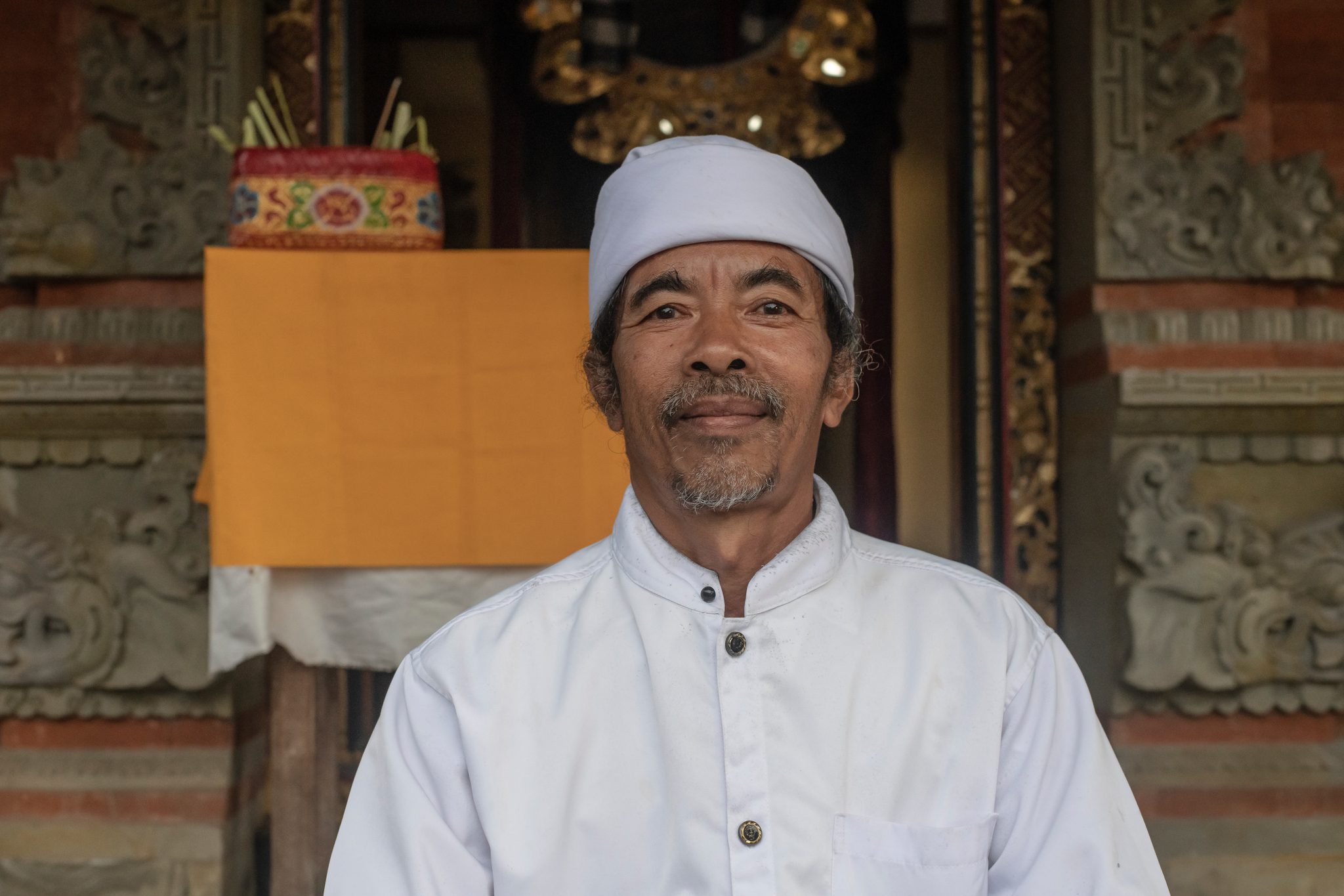 A man in white smiles gently in front of the carved shrine.