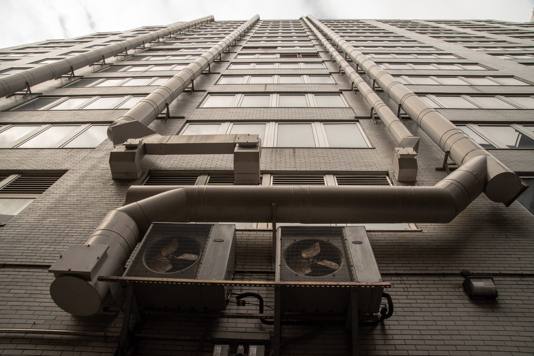 Giant ducts and fans cling to the face of a building, seen straight up from the sidewalk.