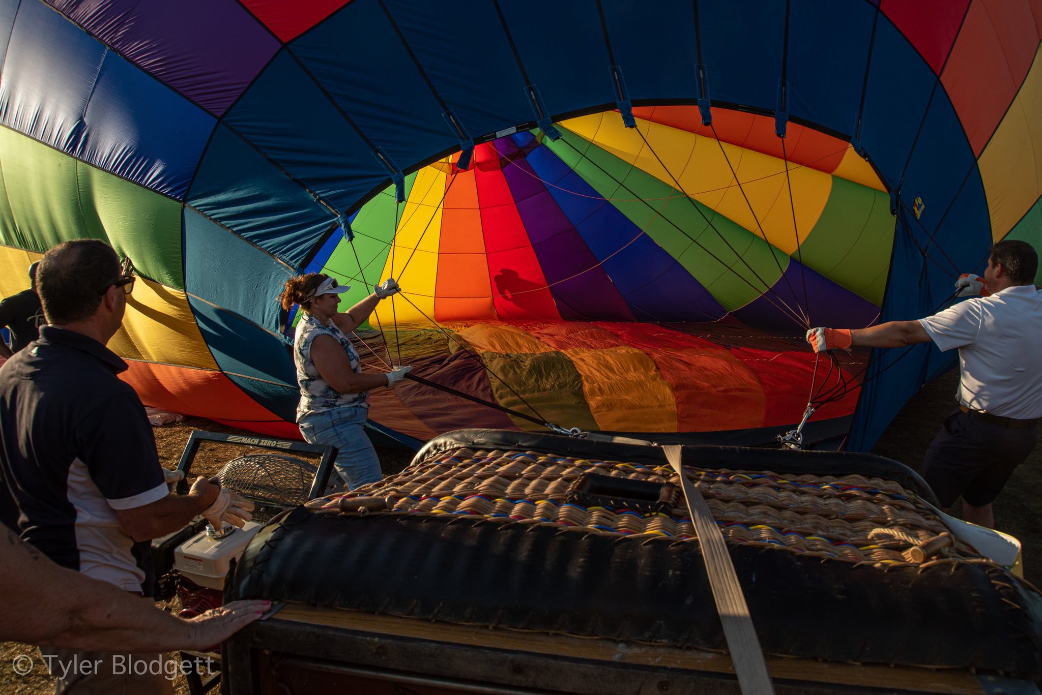 The crew holds open the mouth of a rainbow envelope while the basket waits on its side. Every flight begins like this, held up by many hands.