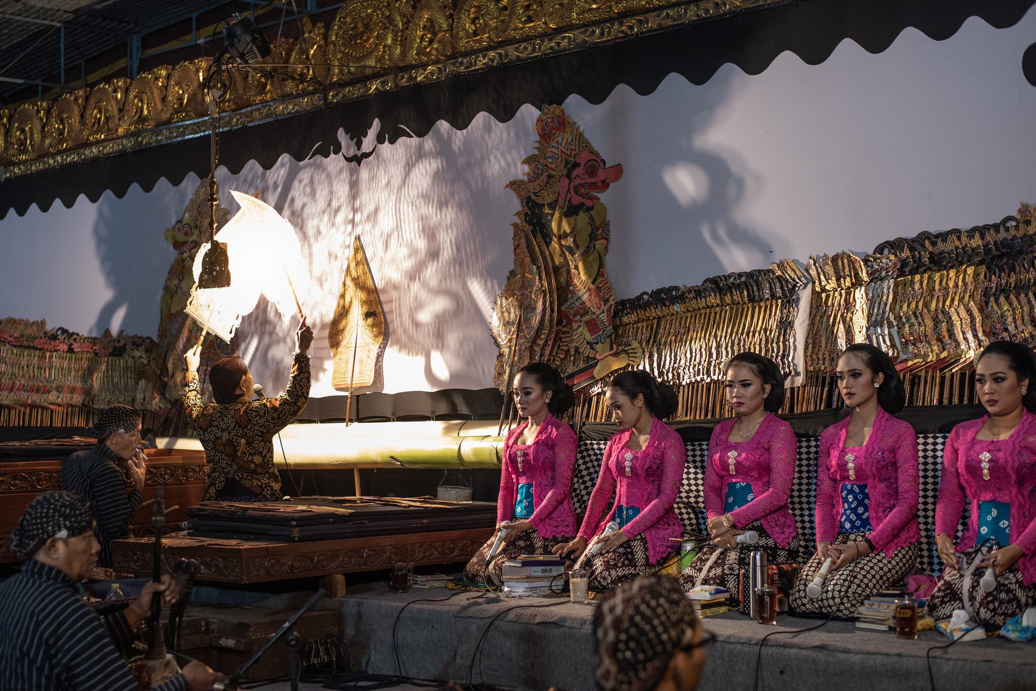 Ki Seno raises a puppet to the lamp while five singers in pink kneel in a row beside him.