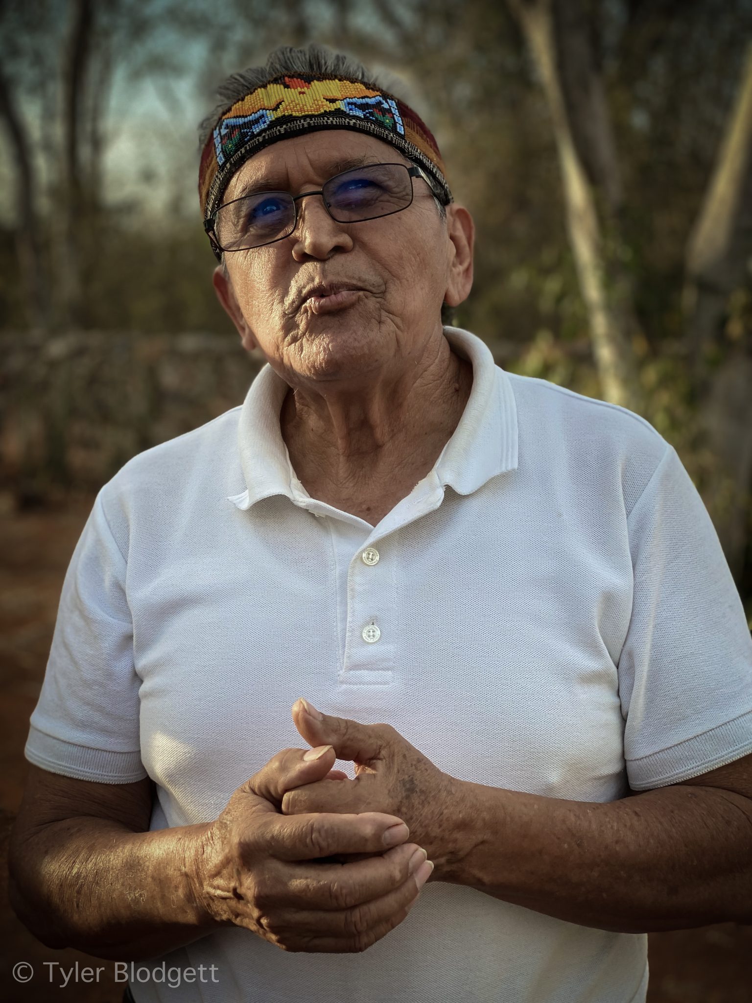 An elder in a beaded headband speaks, hands folded at his chest.